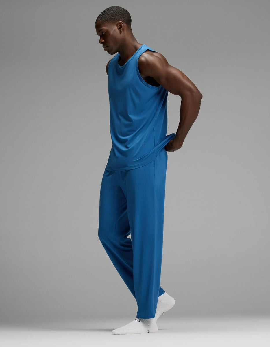Wearing Cozy Earth’s Men’s First Layer Pajama Tank & Pant Set in blue, a person stands against a plain gray background with their hands behind their back, looking down. They complete the look with white socks.