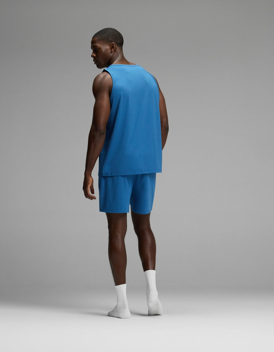 A man stands facing away, dressed in the Cozy Earth Men’s First Layer Pajama Tank & Short Set in blue with white socks, set against a plain gray background.|Color:Anchor