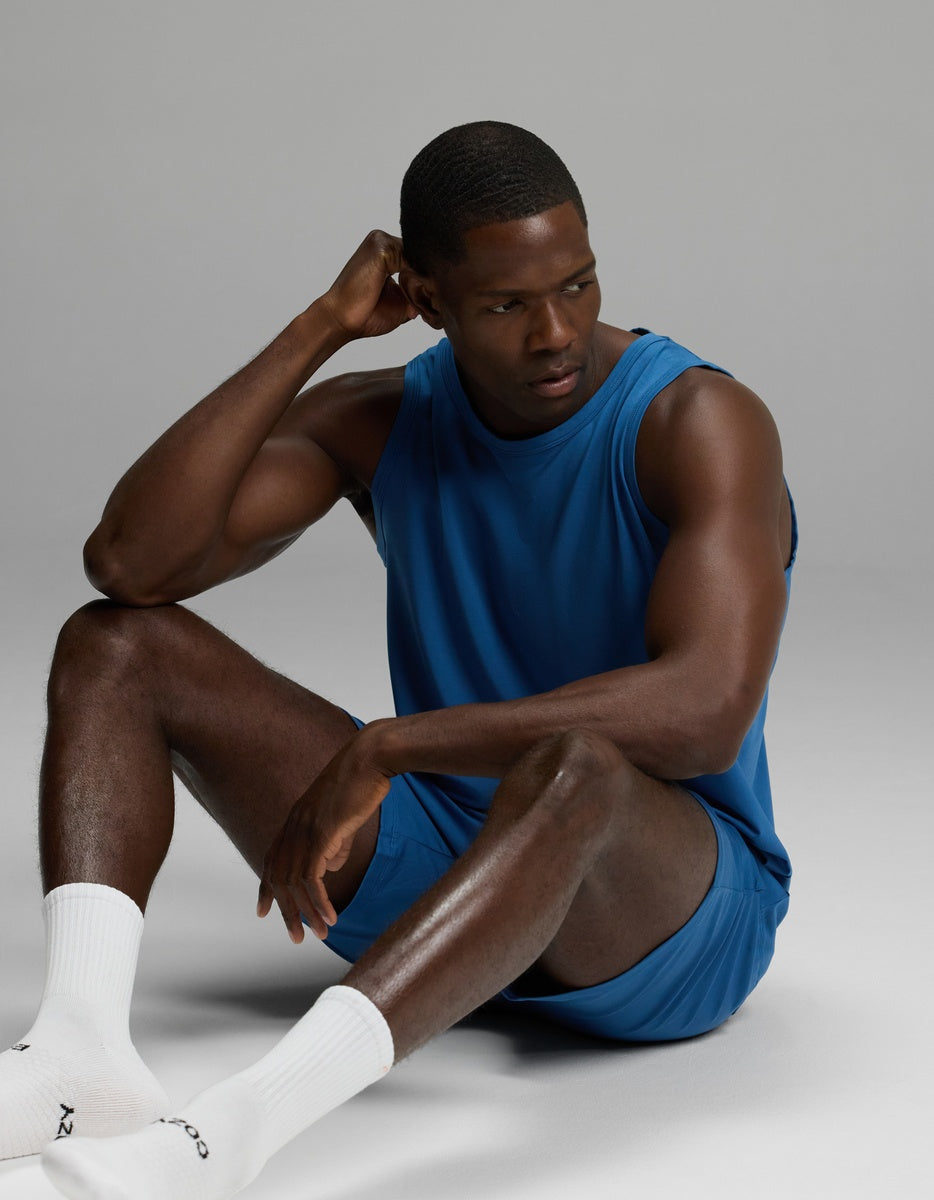 A man models the Cozy Earth Men’s First Layer Pajama Tank & Short Set in blue, sitting on the floor with one hand resting on his head and looking to the side. He completes the look with white socks against a plain, light gray background.
