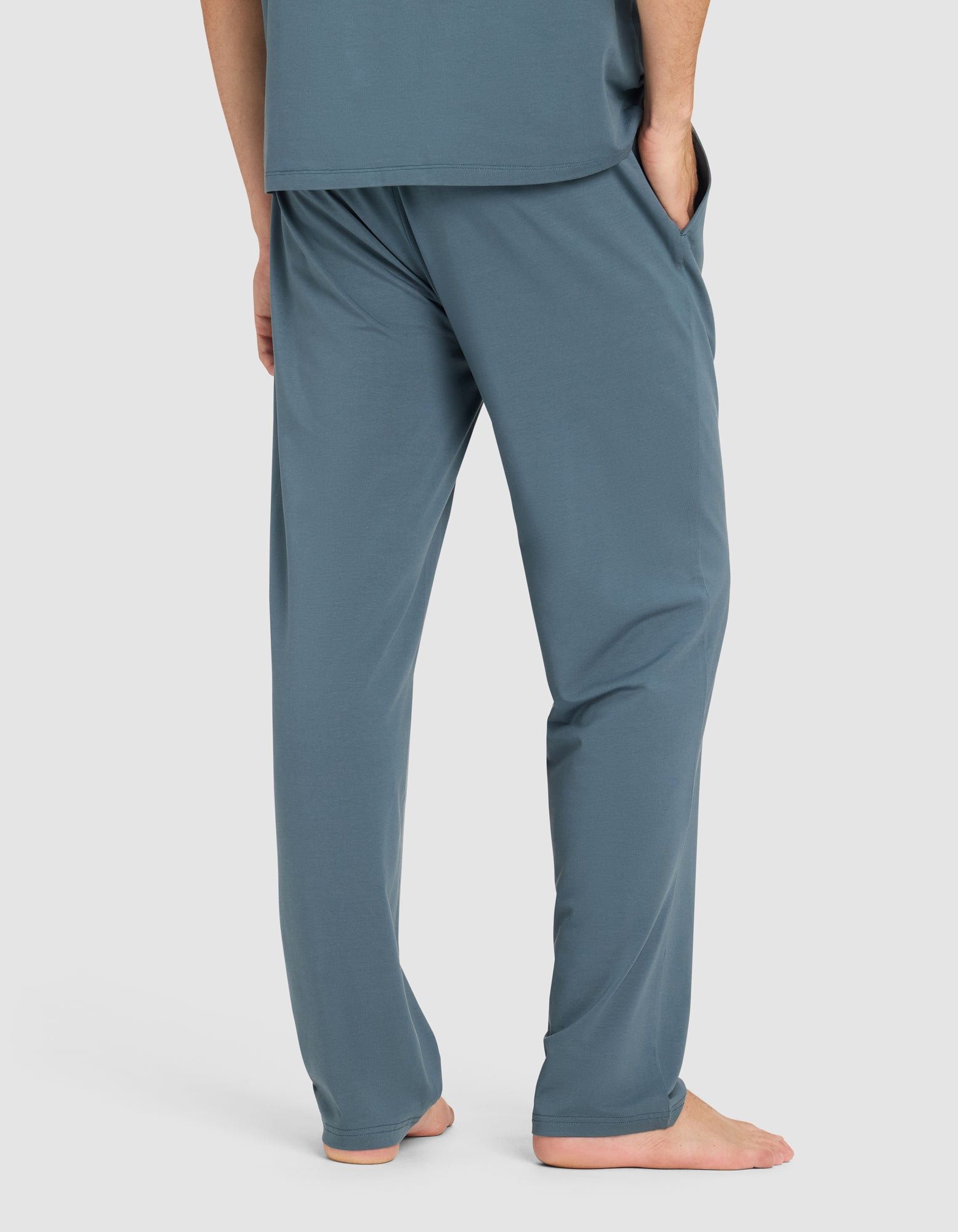 A man stands barefoot with hands in pockets, wearing Cozy Earth's Men's First Layer Pajama Pant in teal blue, paired with a matching top. He is shown from the waist down against a plain light background.
