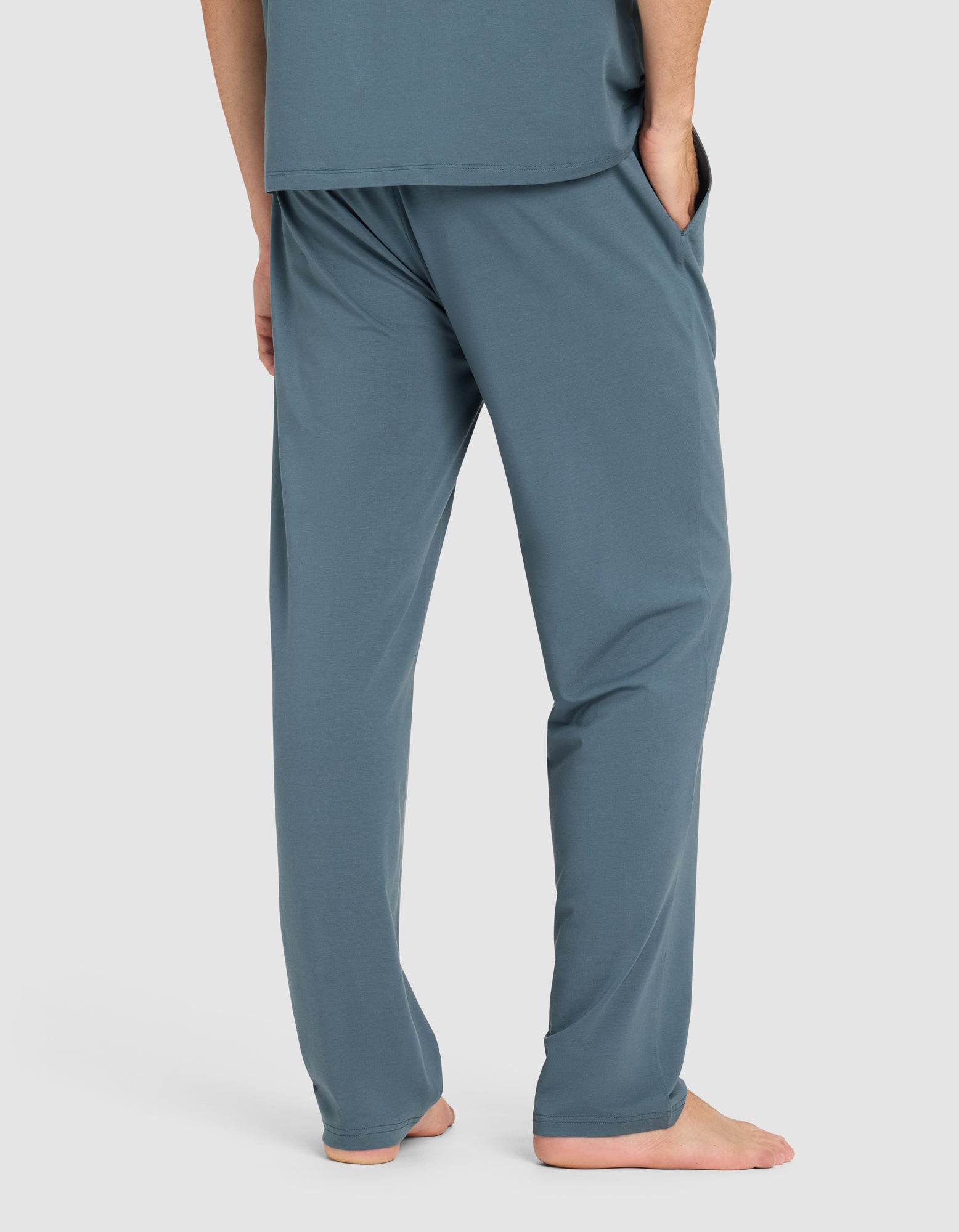 A man stands barefoot with hands in pockets, wearing Cozy Earth's Men's First Layer Pajama Pant in teal blue, paired with a matching top. He is shown from the waist down against a plain light background.