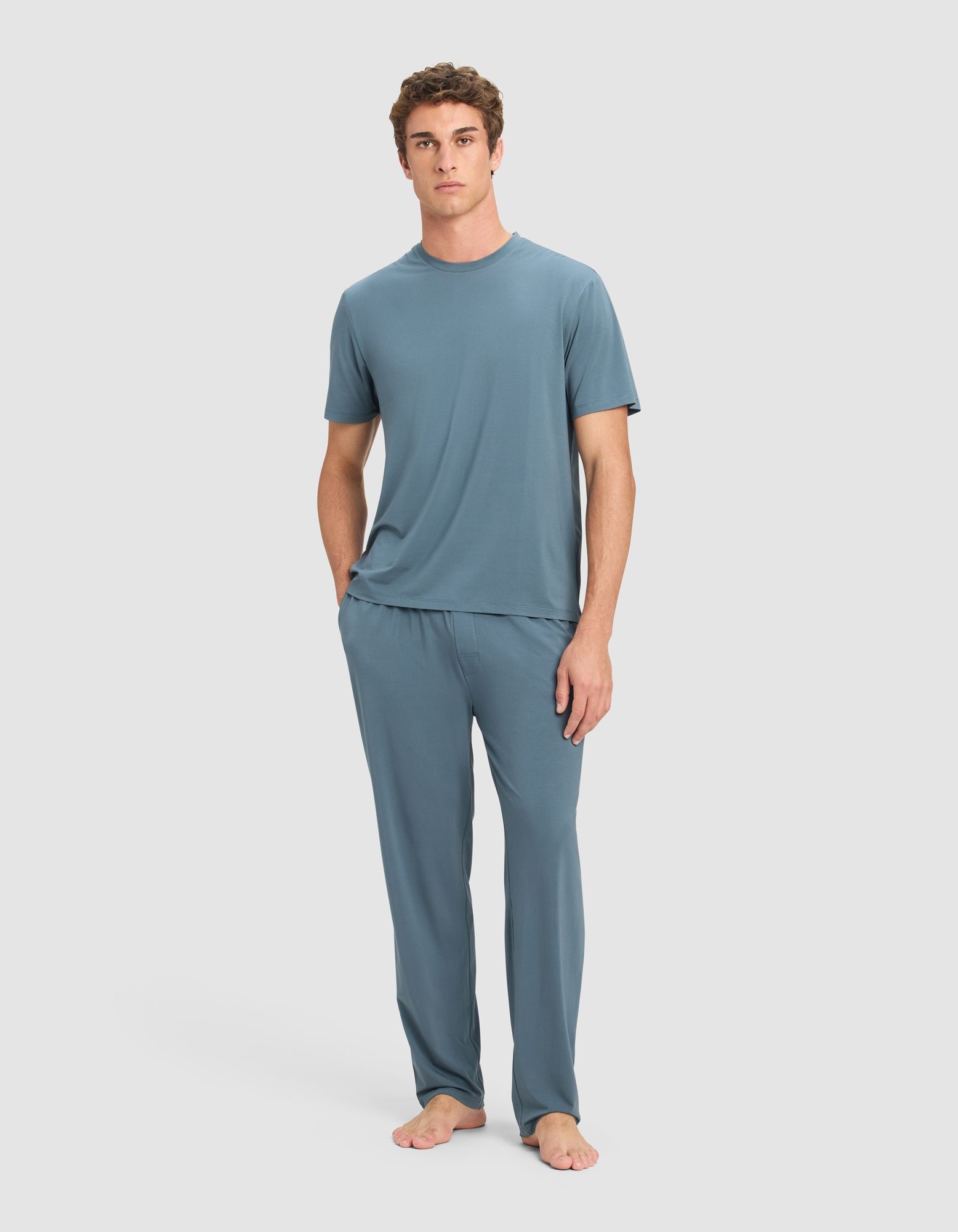 A person stands barefoot against a plain background, wearing Cozy Earth's Men's First Layer Pajama Pant in blue with a matching top. Their left hand is in a pocket, and they face forward with a neutral expression.