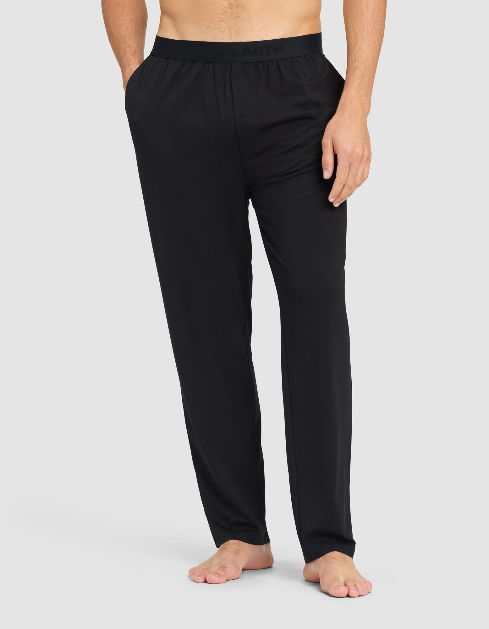 A man, with his upper body and face out of frame, stands barefoot against a light gray background wearing Cozy Earth Men's First Layer Pajama Pant—loose-fitting black pants with an elastic waistband.