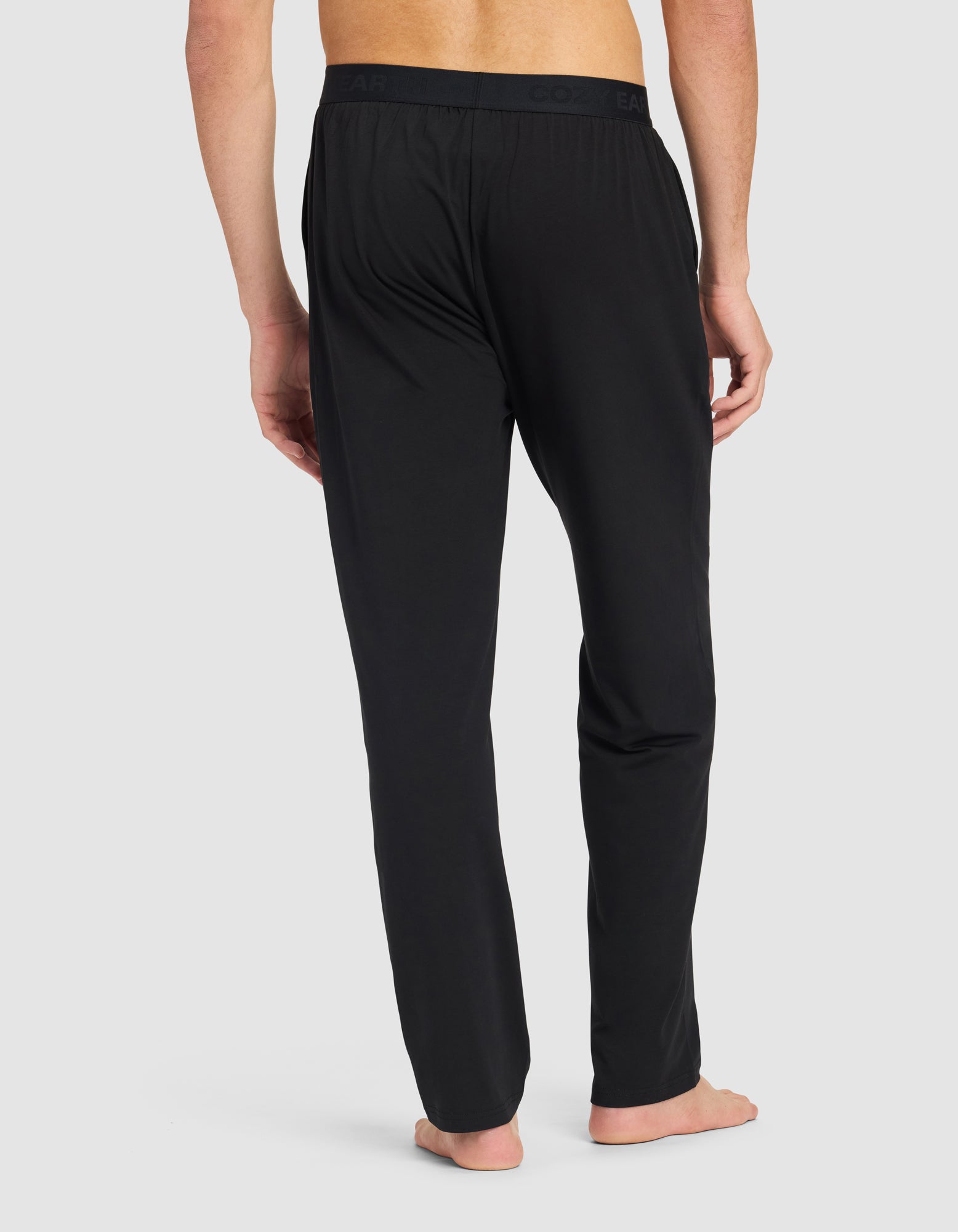 A man stands barefoot, facing away, wearing Cozy Earth Men's First Layer Pajama Pant with an elastic waistband. The background is plain light gray.