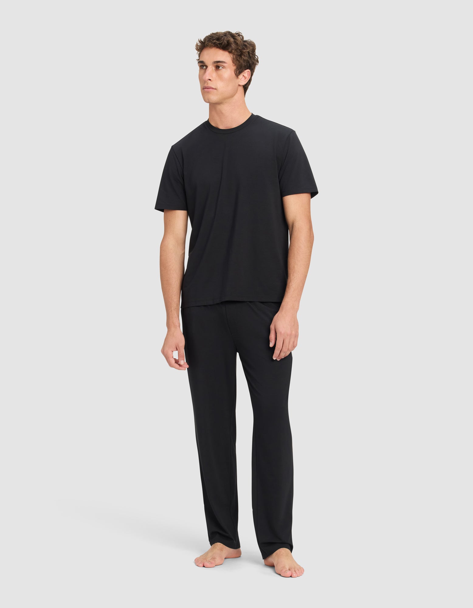 A man stands barefoot against a light background, wearing Cozy Earth's Men's First Layer Pajama Pant in black and a loose-fitting black short-sleeve t-shirt, gazing slightly to the side with a neutral expression.|Color:Jet Black