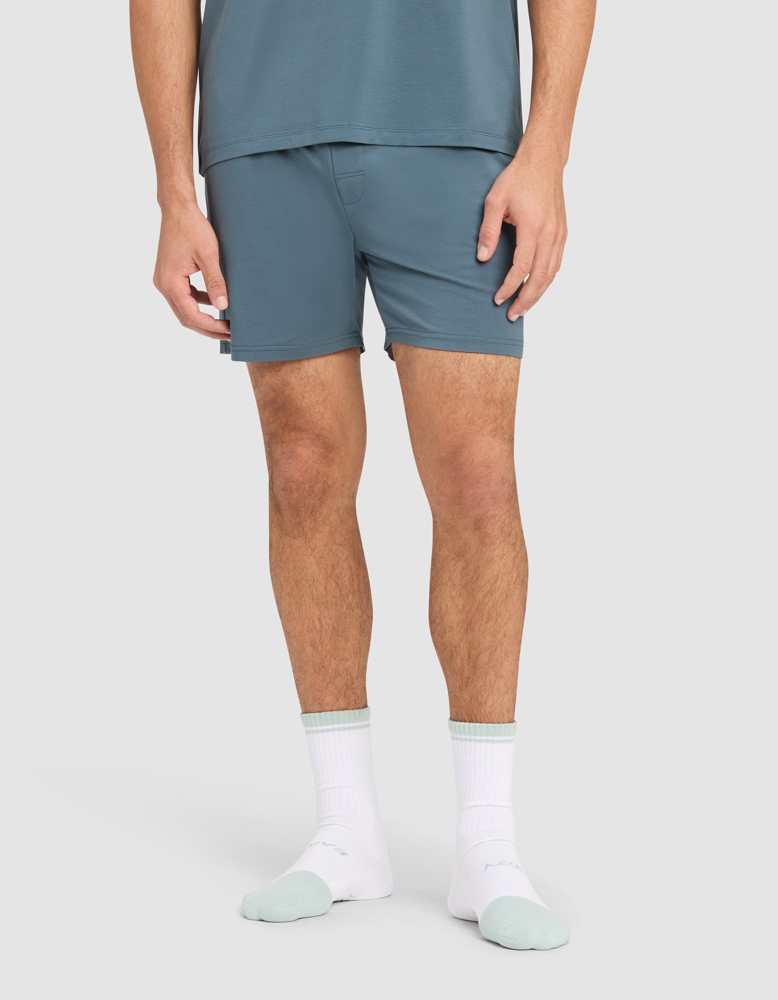 A person stands against a plain background wearing Cozy Earth's Men's First Layer Pajama Short and matching shirt, paired with white socks featuring mint green toes and heels. The image is cropped at the mid-torso. 