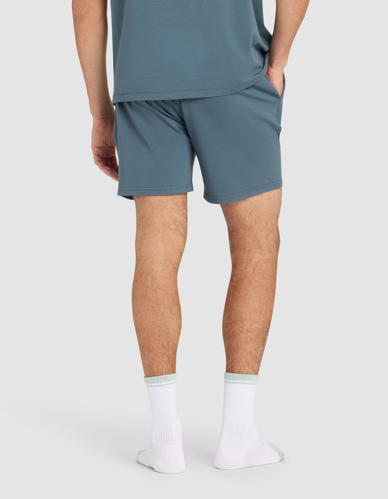A person stands with hands in pockets, wearing Cozy Earth Men's First Layer Pajama Short in blue-green and a matching T-shirt, paired with white socks. The background is plain and light-colored.