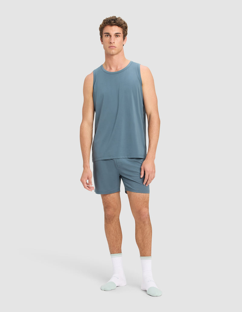 A man stands against a plain background wearing Cozy Earth's Men's First Layer Pajama Shorts and a matching sleeveless blue tank, paired with white socks and light blue slippers. He faces forward with a neutral expression.