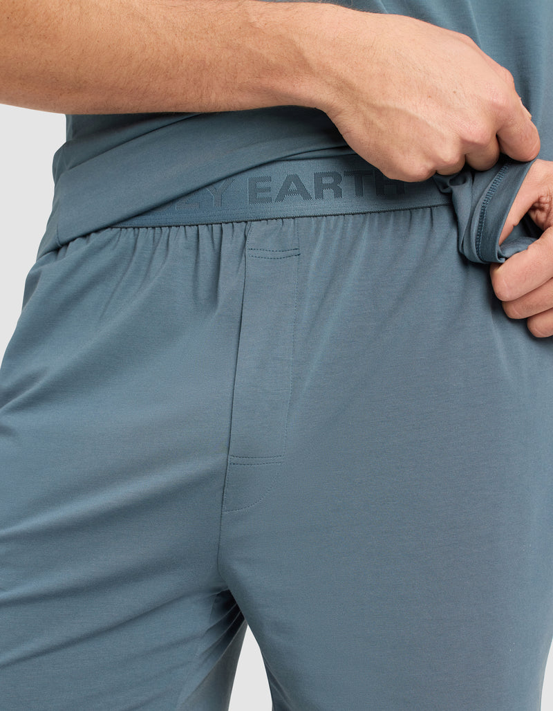 A person wearing Cozy Earth Men's First Layer Pajama Shorts slightly pulls the blue-gray waistband aside, revealing "EARTH" printed on the inner band. Only the lower torso and hand are visible.