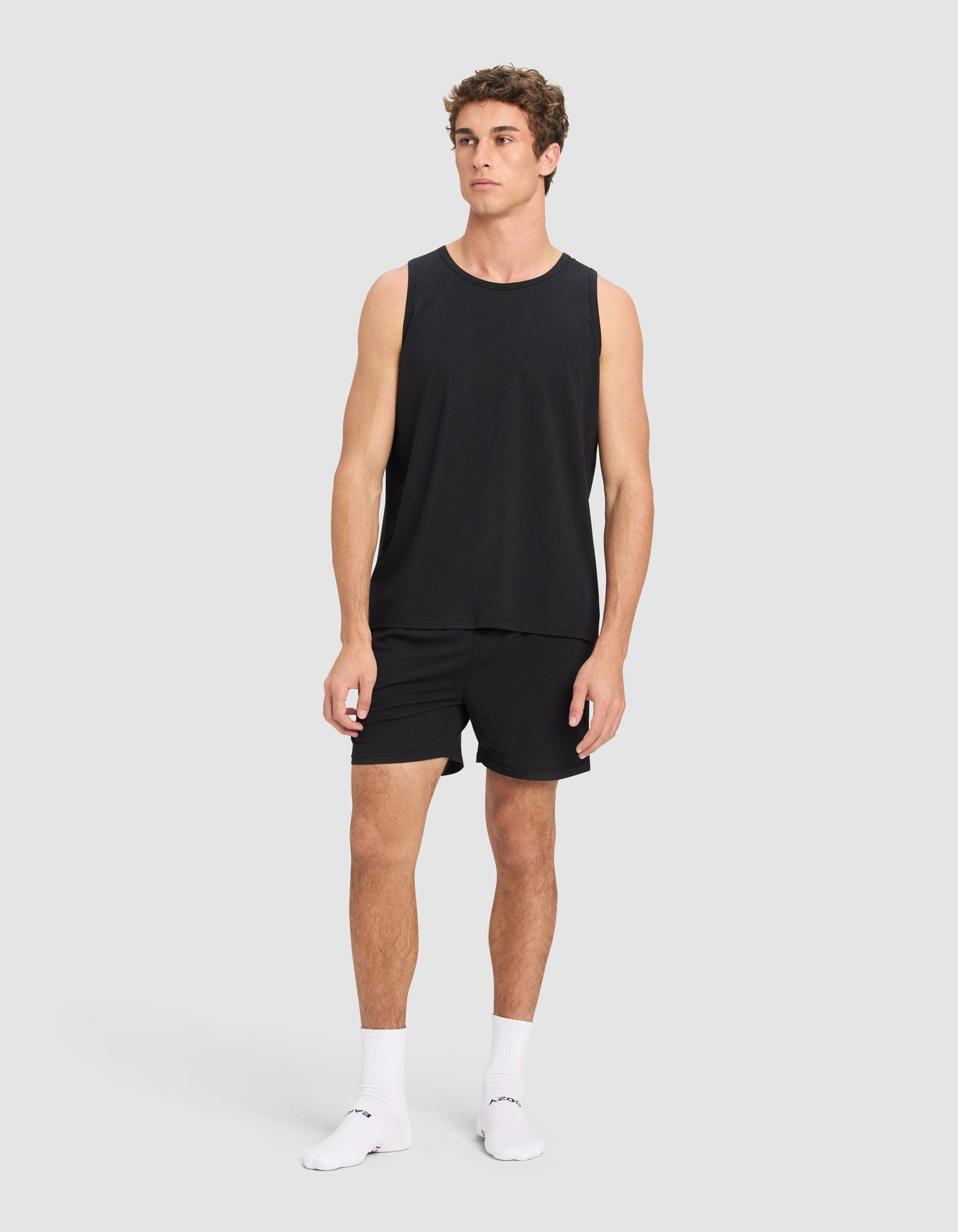 A man stands against a plain light gray background wearing Cozy Earth Men's First Layer Pajama Shorts. He pairs them with a black sleeveless tank top and white ankle socks with black writing, looking slightly to his left with a neutral expression.
