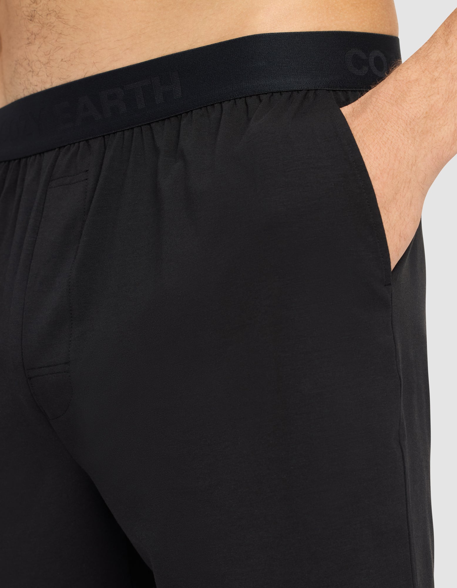 Close-up of a person wearing Cozy Earth Men's First Layer Pajama Short in black, featuring a subtle-lettered waistband and one hand in the pocket. The background is plain and light-colored.