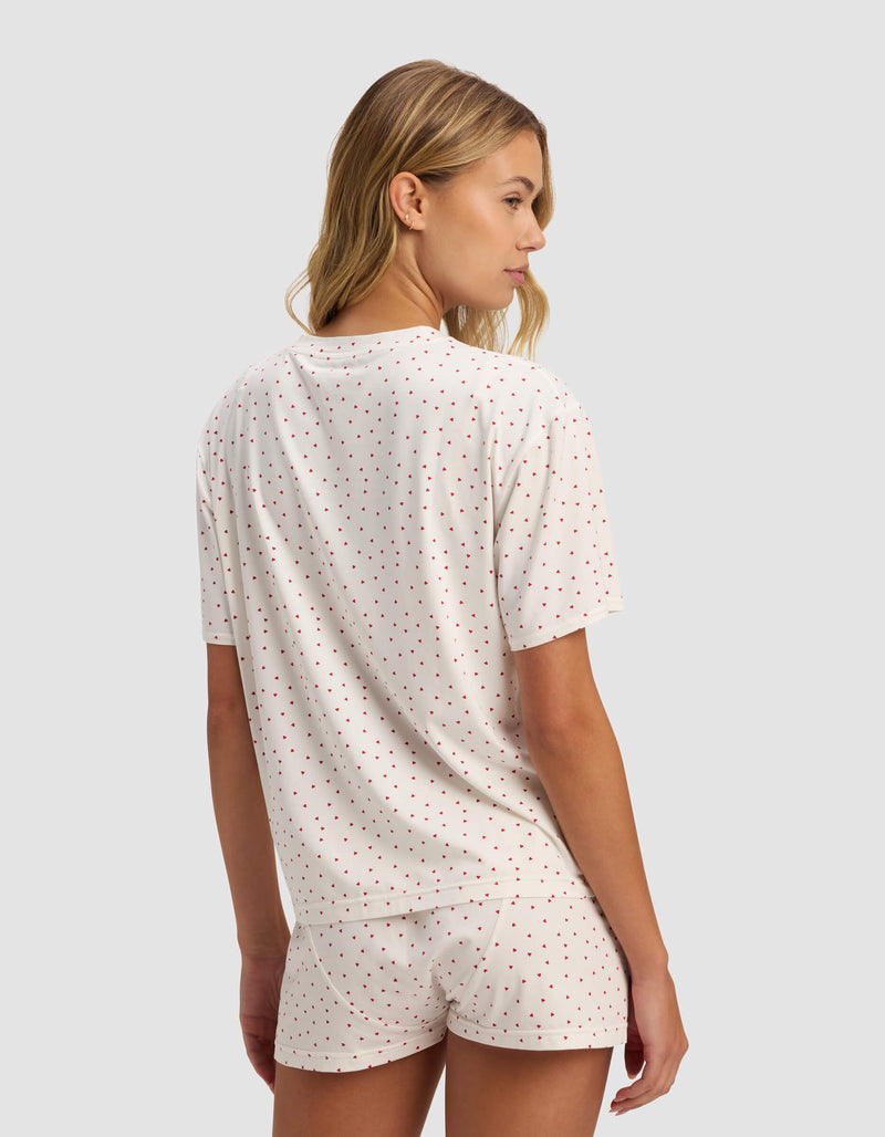 A woman with wavy blonde hair stands slightly turned away, wearing the Cozy Earth Women's First Layer Pajama Short Sleeve Tee and matching shorts in white with small red polka dots, set against a plain light background.