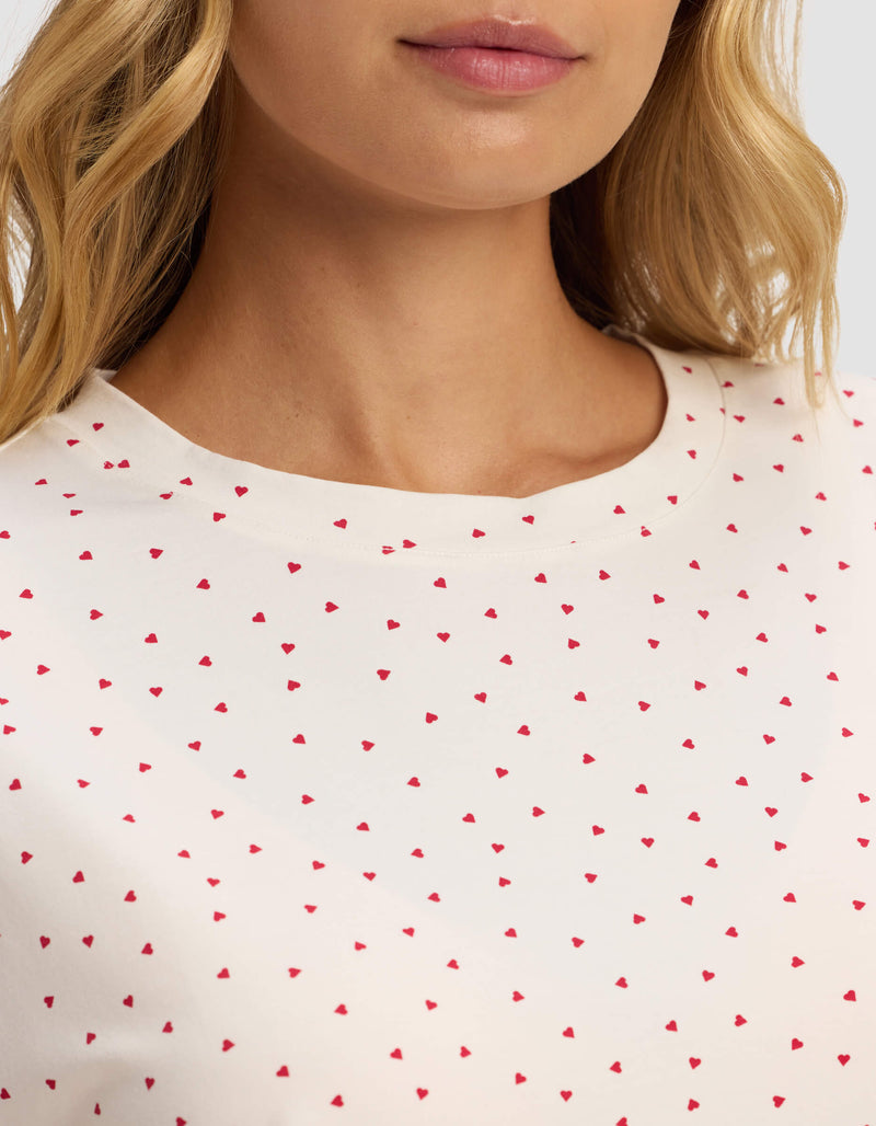 A close-up of a woman wearing the Cozy Earth Women's First Layer Pajama Short Sleeve Tee in white with small red hearts, showing her neck, lower face, and some blonde hair.