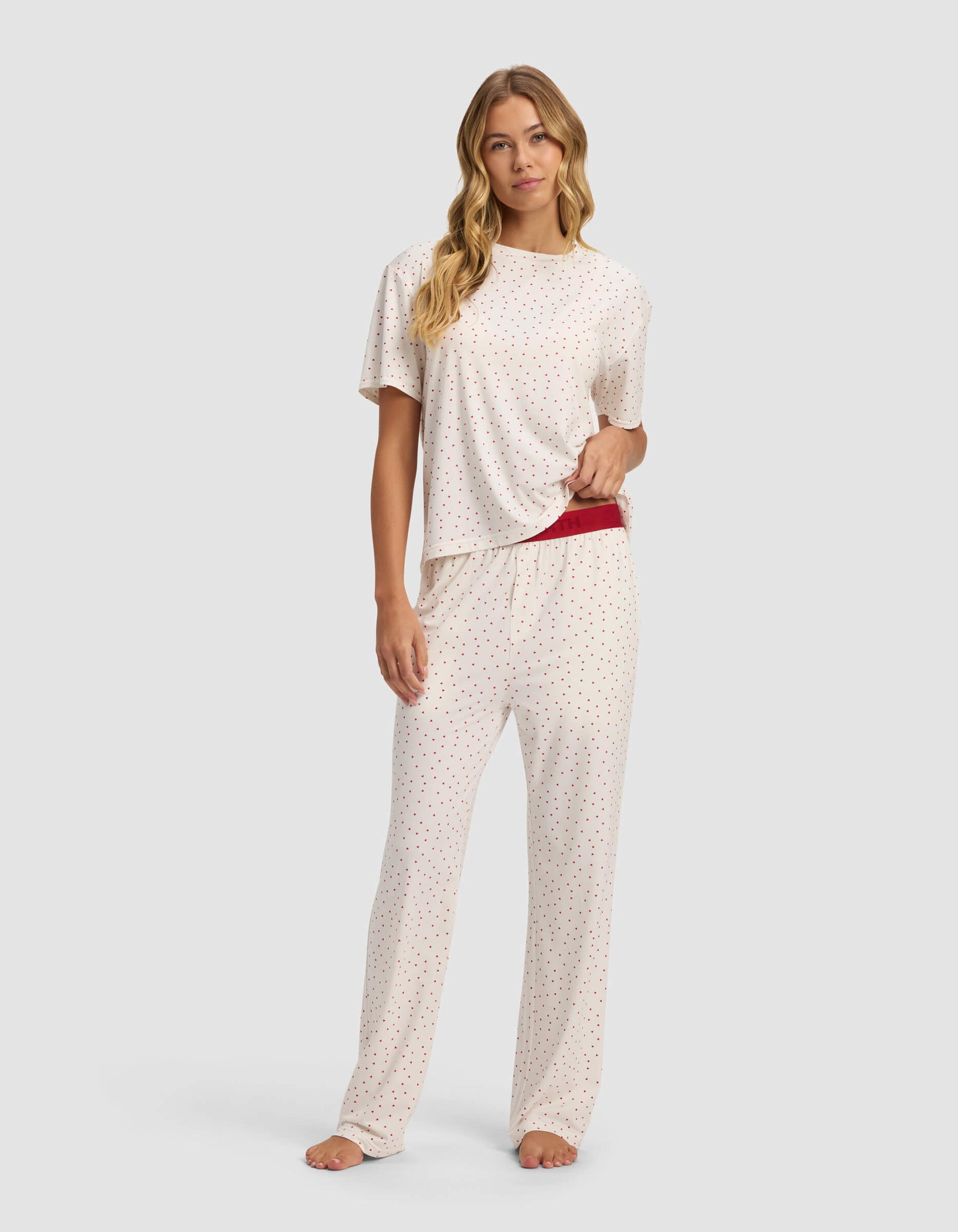 A woman stands barefoot in the Cozy Earth Women's First Layer Pajama Short Sleeve Tee & Pant Set, featuring a relaxed cream color with small polka dots, a red waistband, and long wavy blonde hair.