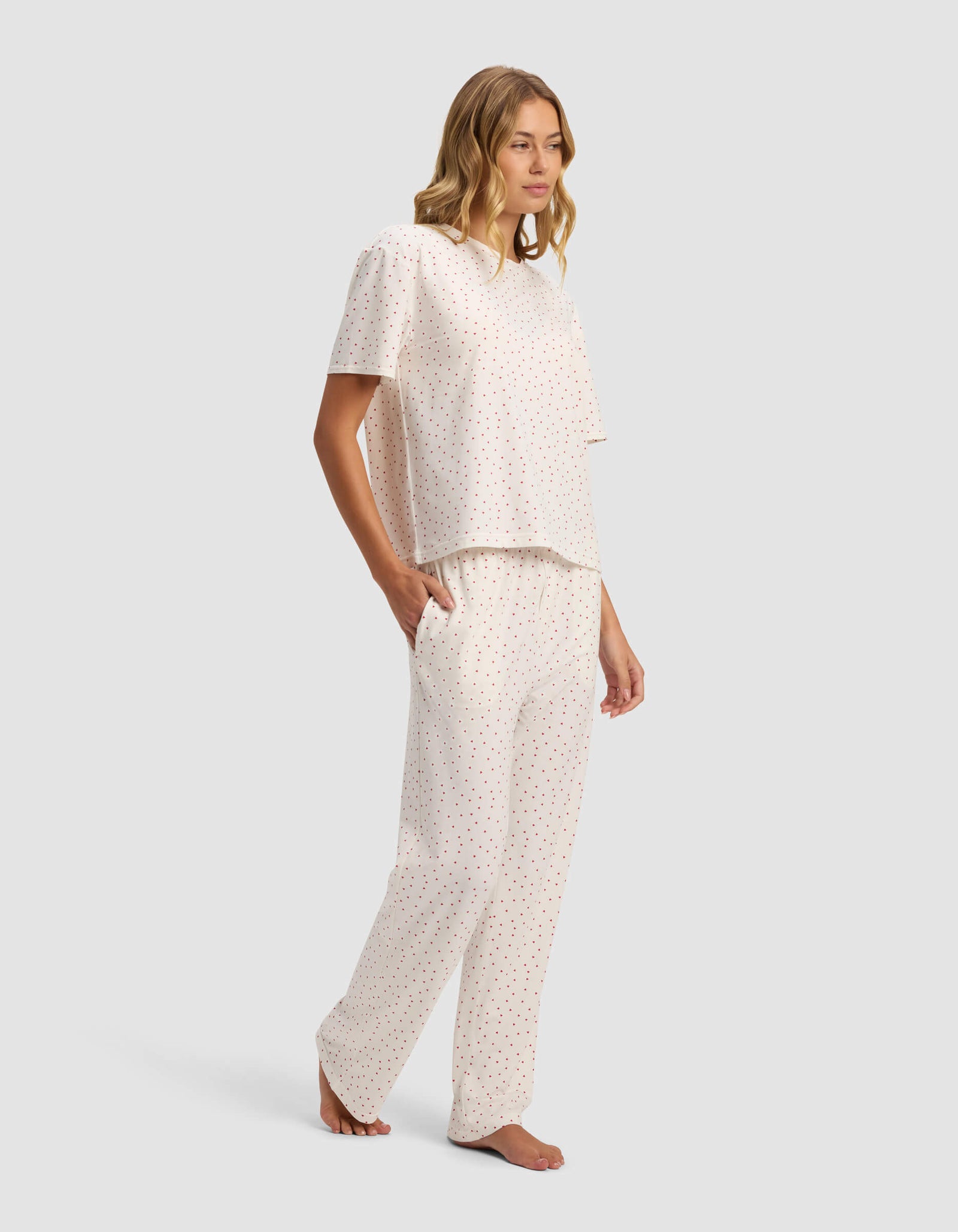 A woman with wavy, shoulder-length hair stands barefoot, wearing Cozy Earth's Women's First Layer Pajama Short Sleeve Tee & Pant Set in white with small multicolored polka dots, facing slightly to the side.
