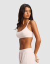 A woman with long dark hair stands in profile, wearing Cozy Earth's Women's First Layer Pajama Soft Bra in light pink with matching shorts, against a plain light gray background.|Color:Ballet
