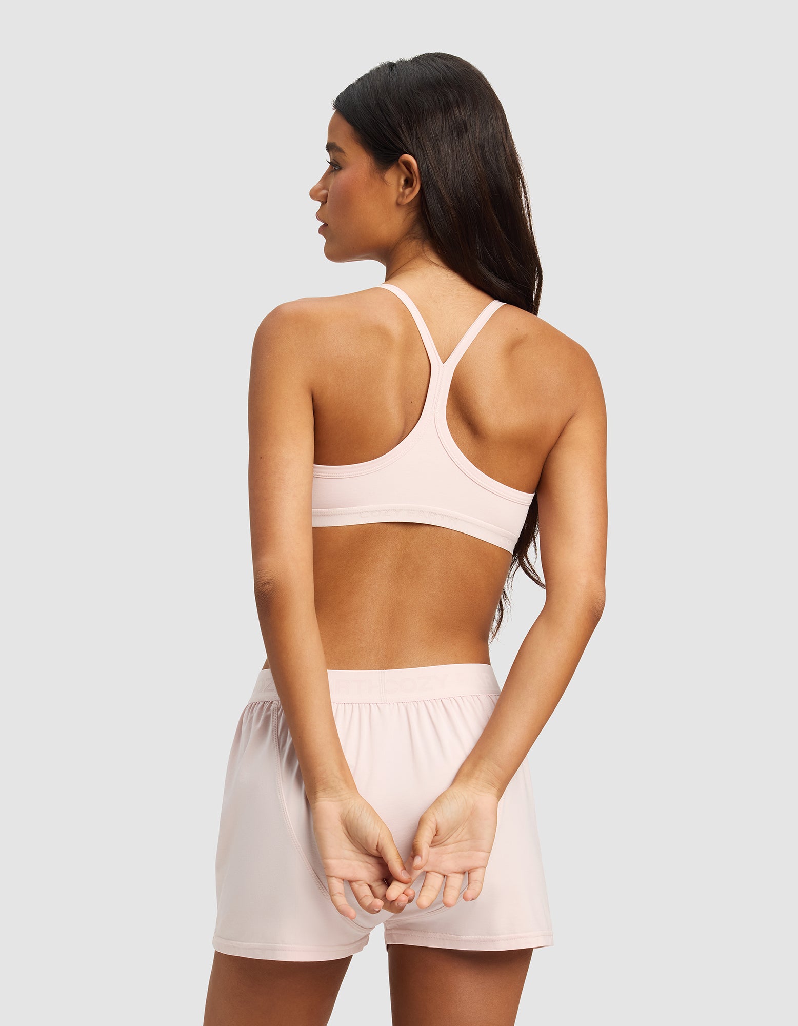 A woman with long dark hair models the Cozy Earth Women's First Layer Pajama Soft Bra in light pink, seen from behind with hands clasped, standing against a plain gray background.