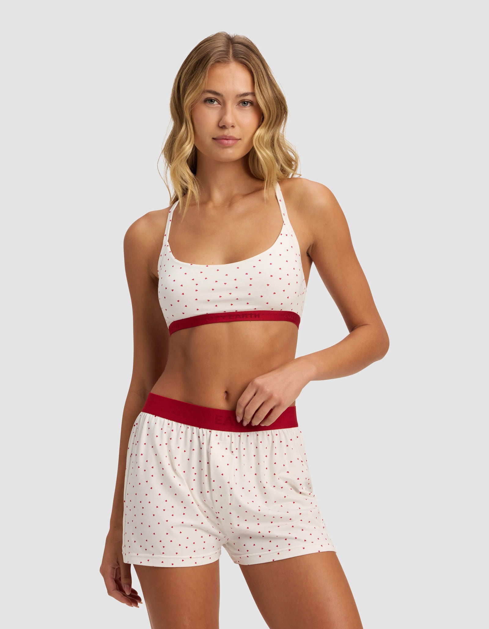 A woman models the Cozy Earth Women's First Layer Pajama Soft Bra set, featuring a white base with small red polka dots, red waistband on the shorts, and a red band on the bralette, against a light gray background.