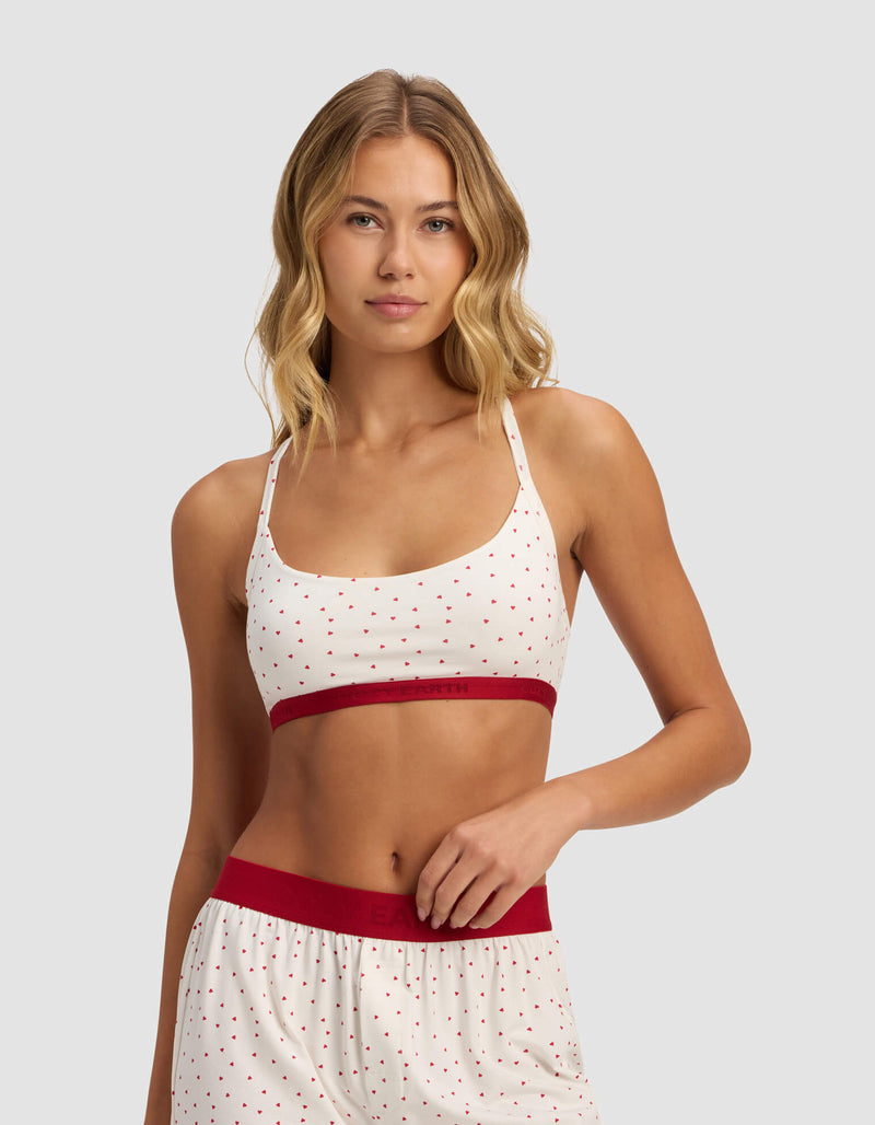 A woman models the Cozy Earth Women's First Layer Pajama Soft Bra set, featuring a white base with small red polka dots, red waistband on the shorts, and a red band on the bralette, against a light gray background.
