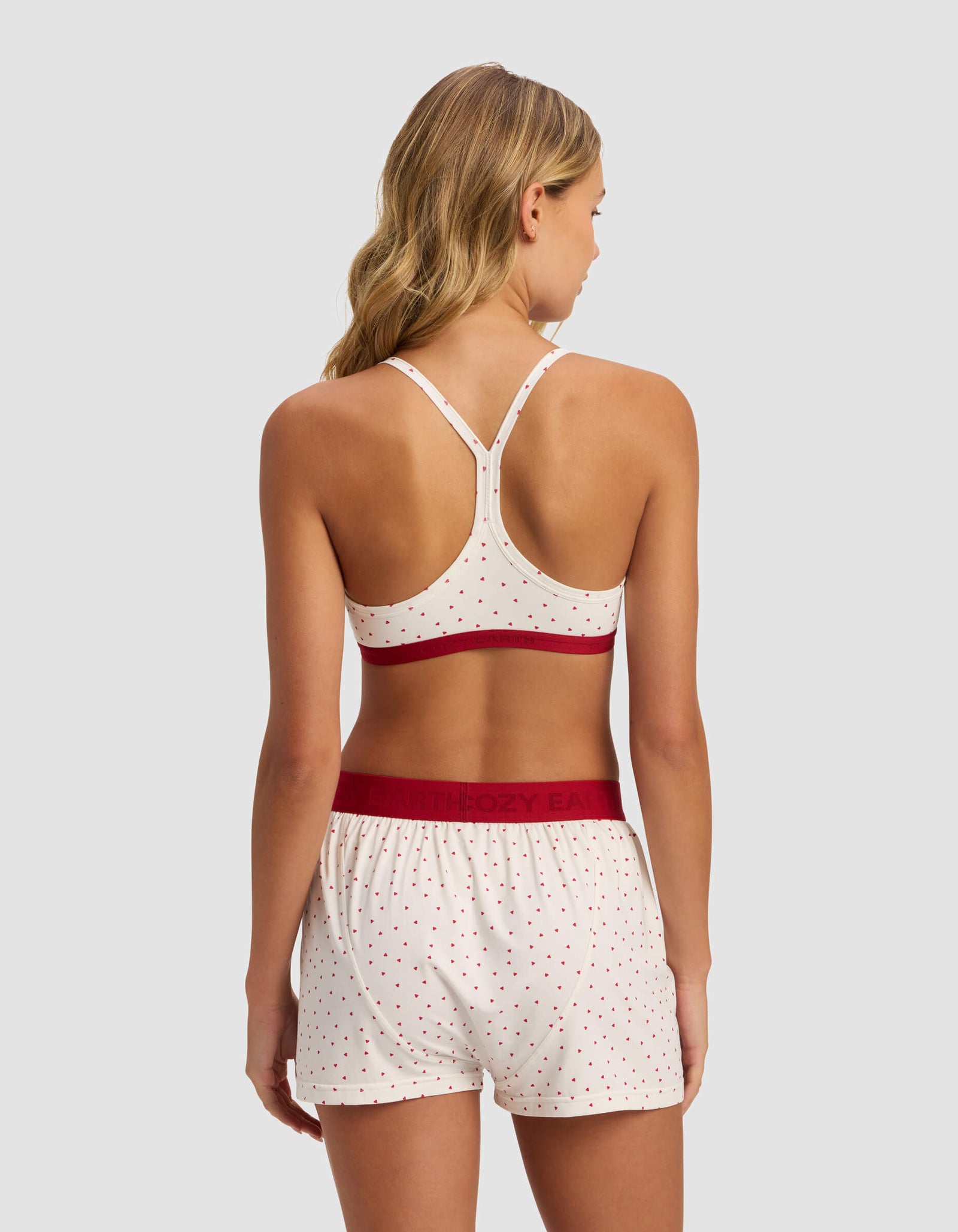 A woman with wavy blonde hair faces away, wearing Cozy Earth's Women's First Layer Pajama Soft Bra and matching shorts in cream with red polka dots, featuring solid red waistband and trim. The background is plain light gray.