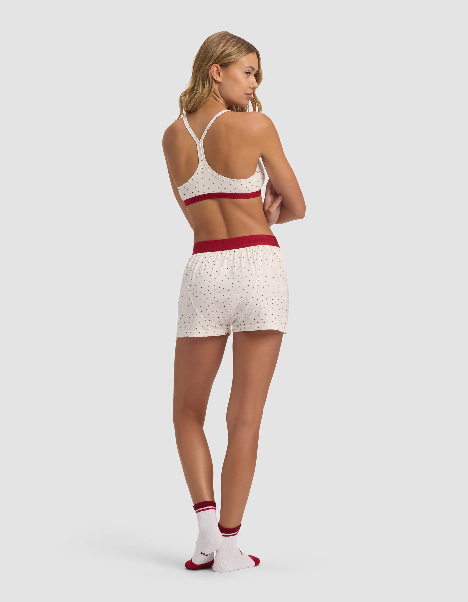 A woman faces away with arms crossed, wearing Cozy Earth's Women's First Layer Pajama Soft Bra & Boxer Short Set—featuring a white racerback tank, white boxer shorts with small red dots and a red waistband, plus matching socks with red trim on a light gray background.|Color:Cranberry Sea Salt Mini Heart