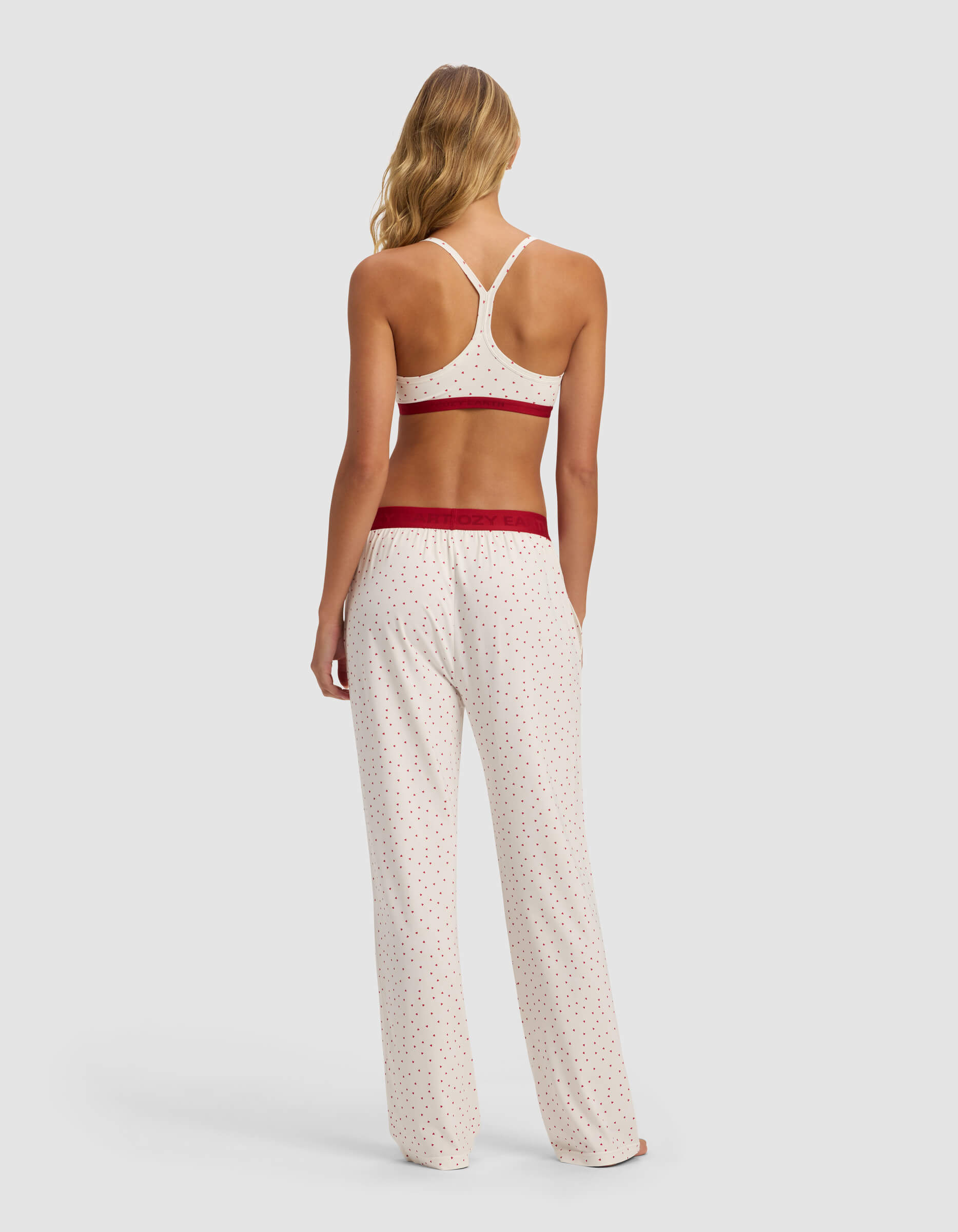 A woman faces away, modeling the Cozy Earth Women's First Layer Pajama Soft Bra & Pant Set: a white racerback bralette and loose pants with red polka dots and waistband, offering a relaxed fit against a plain background.