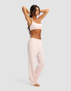 A woman wears the Cozy Earth Women's First Layer Pajama Soft Bra & Pant Set in pale pink, standing barefoot with arms raised, holding her hair back. She faces slightly left against a plain light gray background.|Color:Ballet