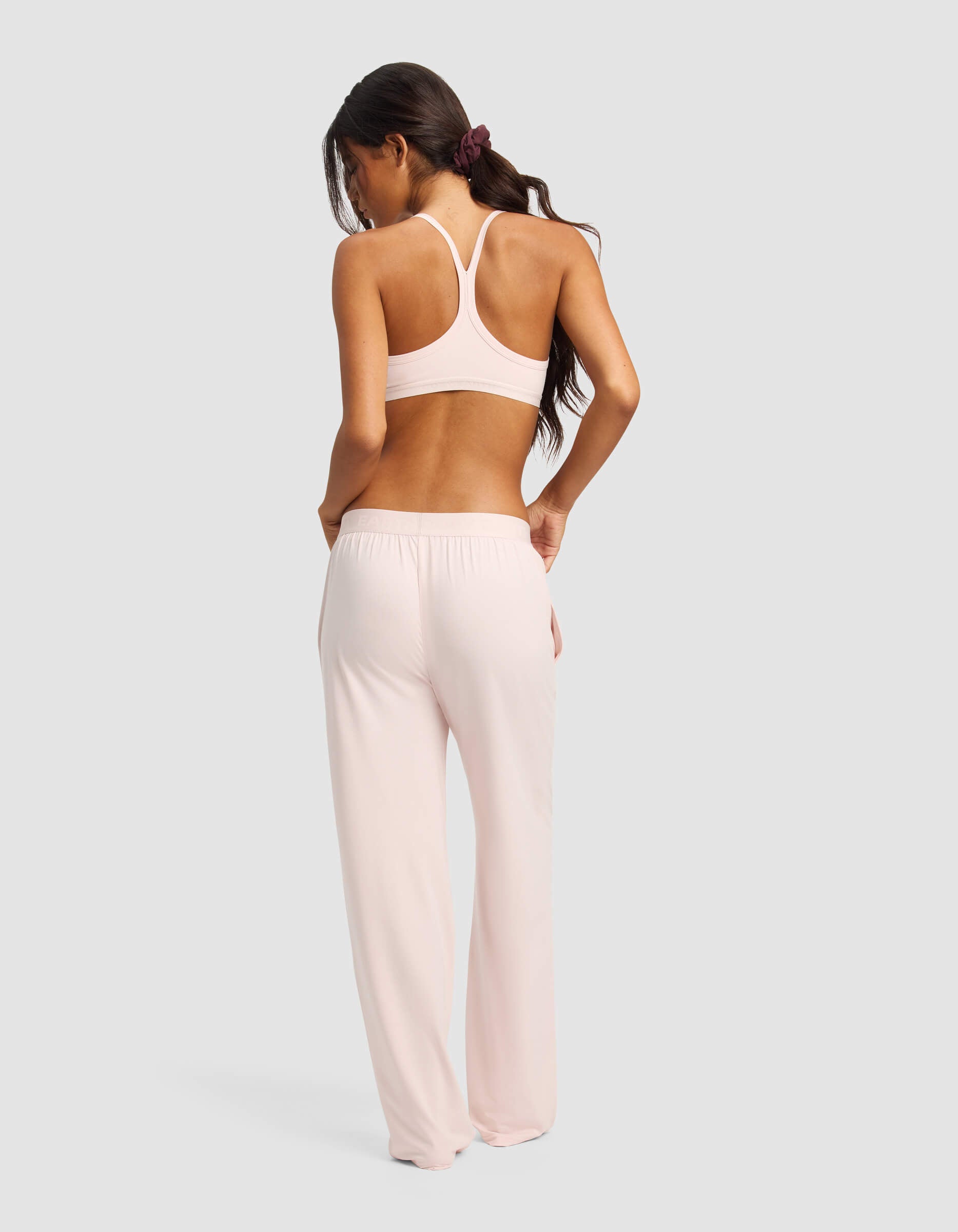 A woman with long dark hair in a ponytail wears Cozy Earth's Women's First Layer Pajama Soft Bra & Pant Set in light pink, standing with her back to a plain light gray background.|Color:Ballet