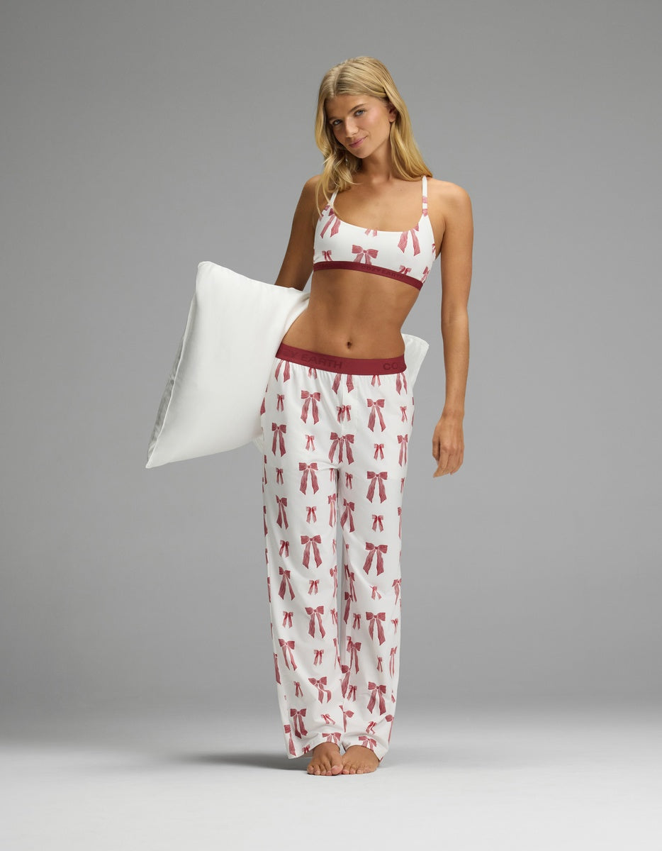 A woman wearing a white Cozy Earth Women’s First Layer Pajama Soft Bra & Pajama Pant Set with a red bow print, holding a white pillow.