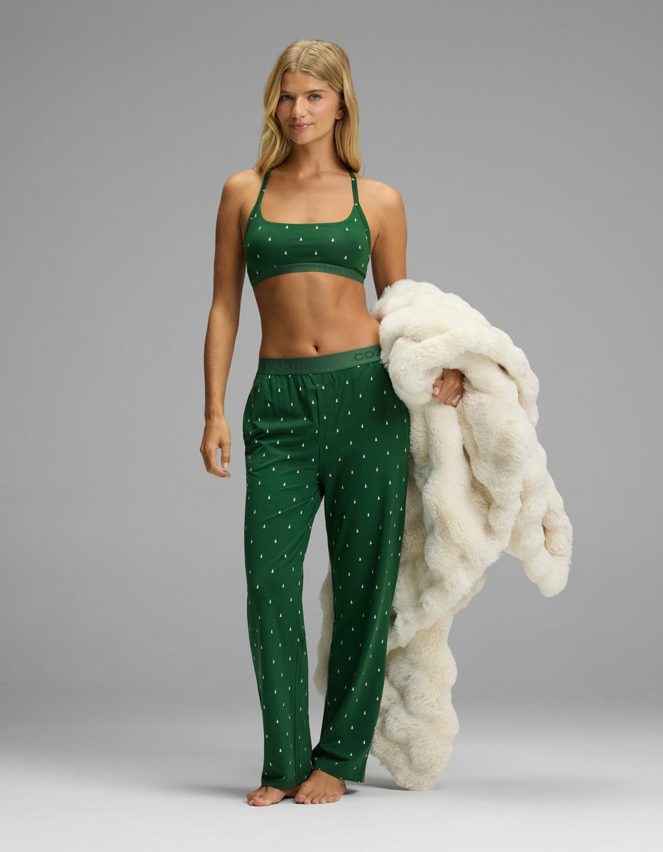 A woman wears a Cozy Earth Women’s First Layer Pajama Soft Bra & Pajama Pant Set in dark green with a white tree pattern while holding a cream-colored faux fur throw blanket.
