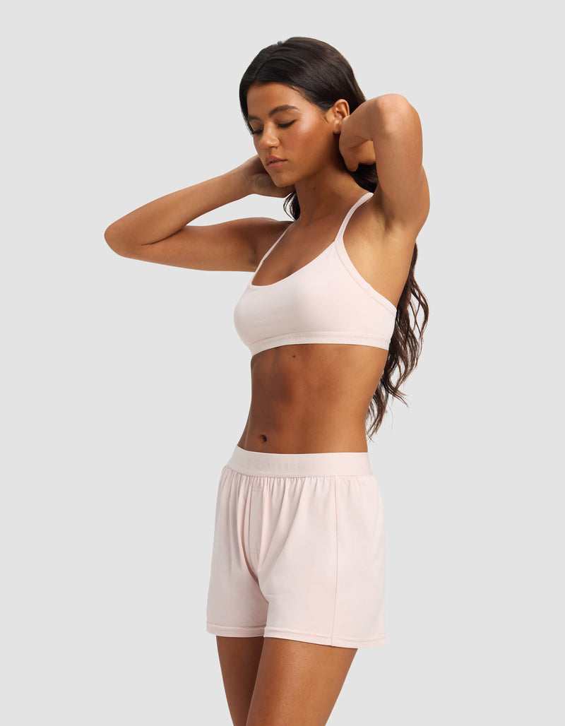 A woman with long dark hair wears the Cozy Earth Women's First Layer Pajama Soft Bra & Boxer Short Set in light pink, standing against a plain gray background and looking down with her hands raised to her head.