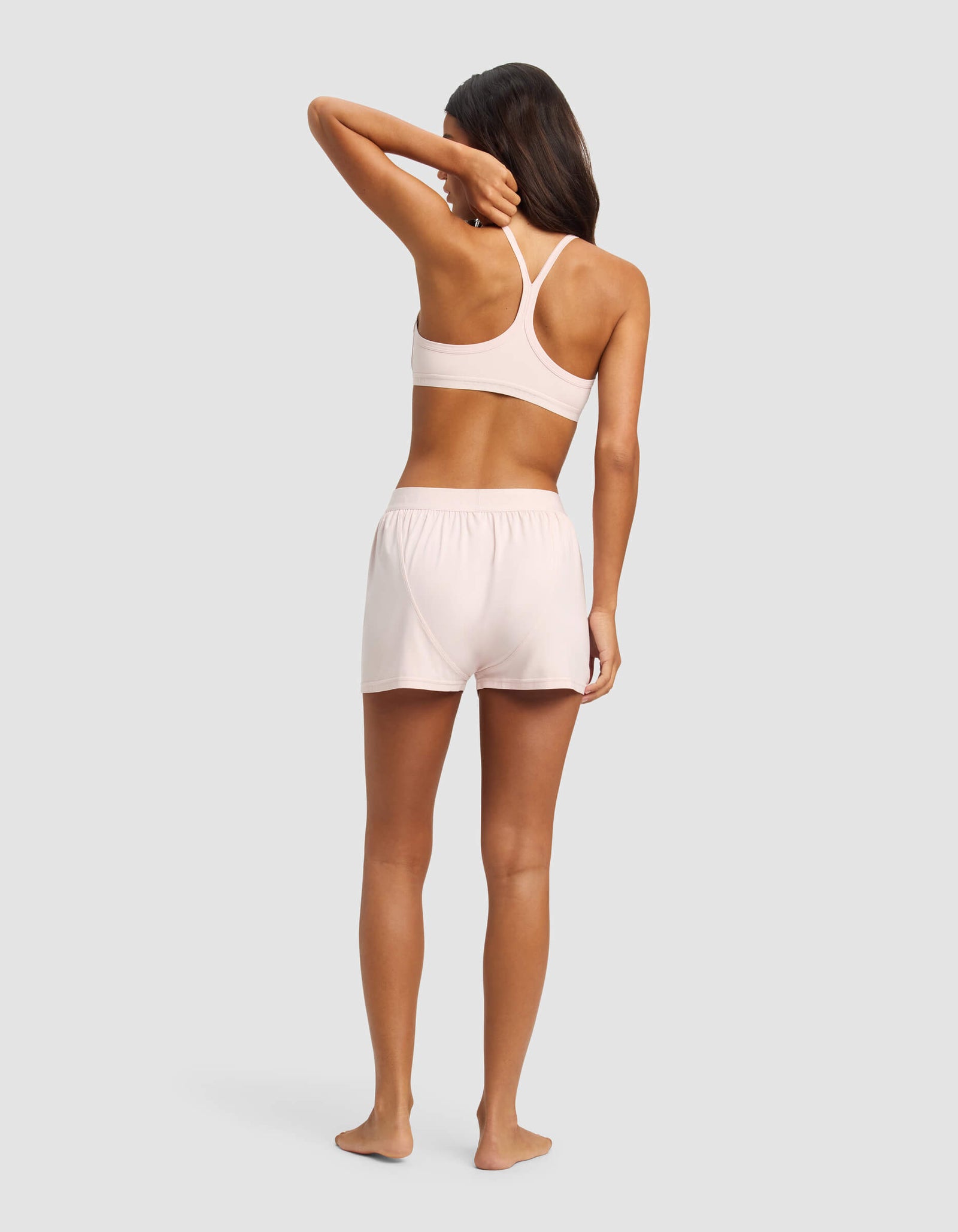 A woman stands barefoot on a gray background, wearing the Cozy Earth Women's First Layer Pajama Soft Bra & Boxer Short Set in light pink. She faces away, with one arm bent and her hand touching her head.
