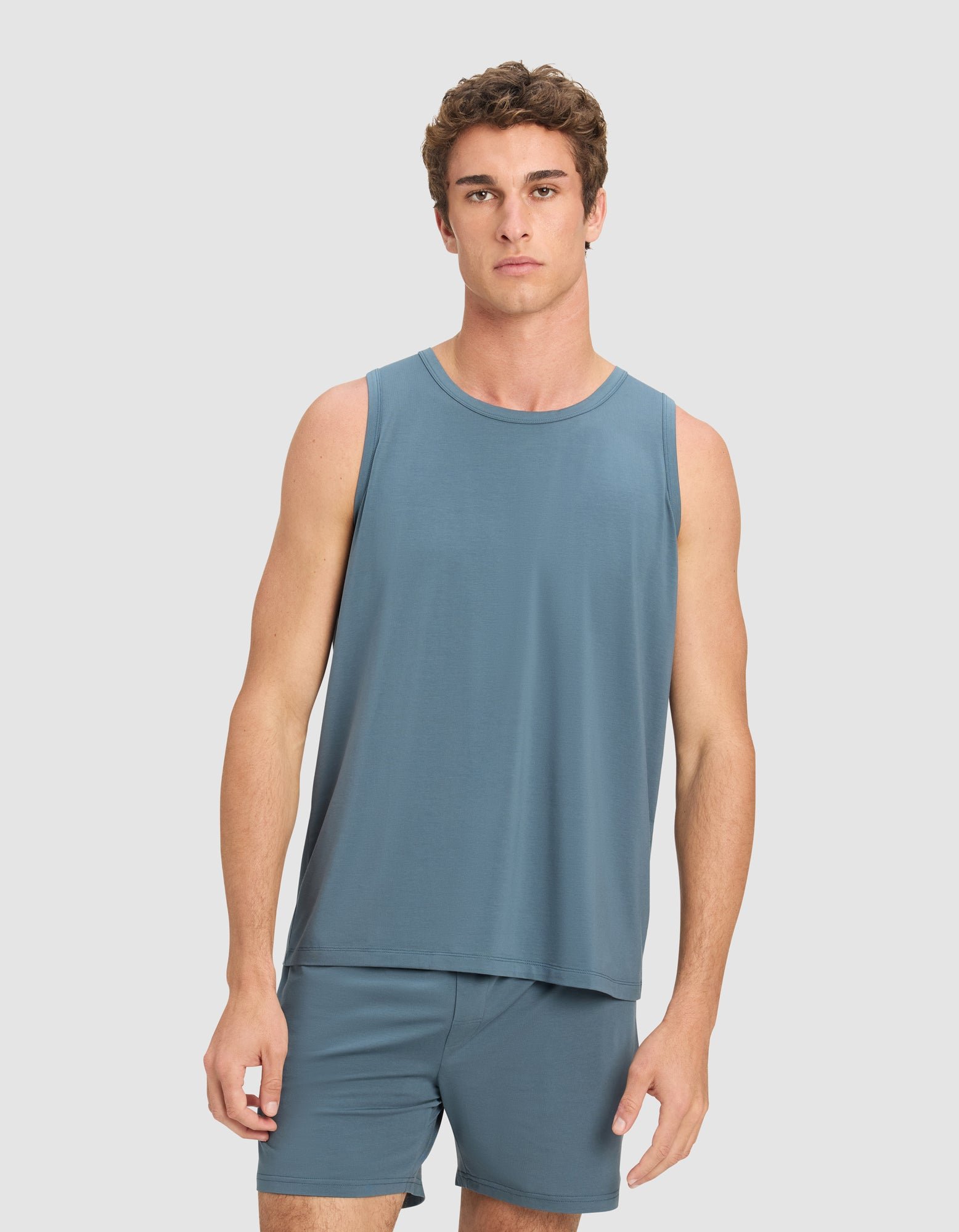 A young man with short, curly brown hair wears the Cozy Earth Men's First Layer Pajama Tank in blue, paired with matching shorts, standing against a plain light gray background and looking directly at the camera.|Color:Dawn