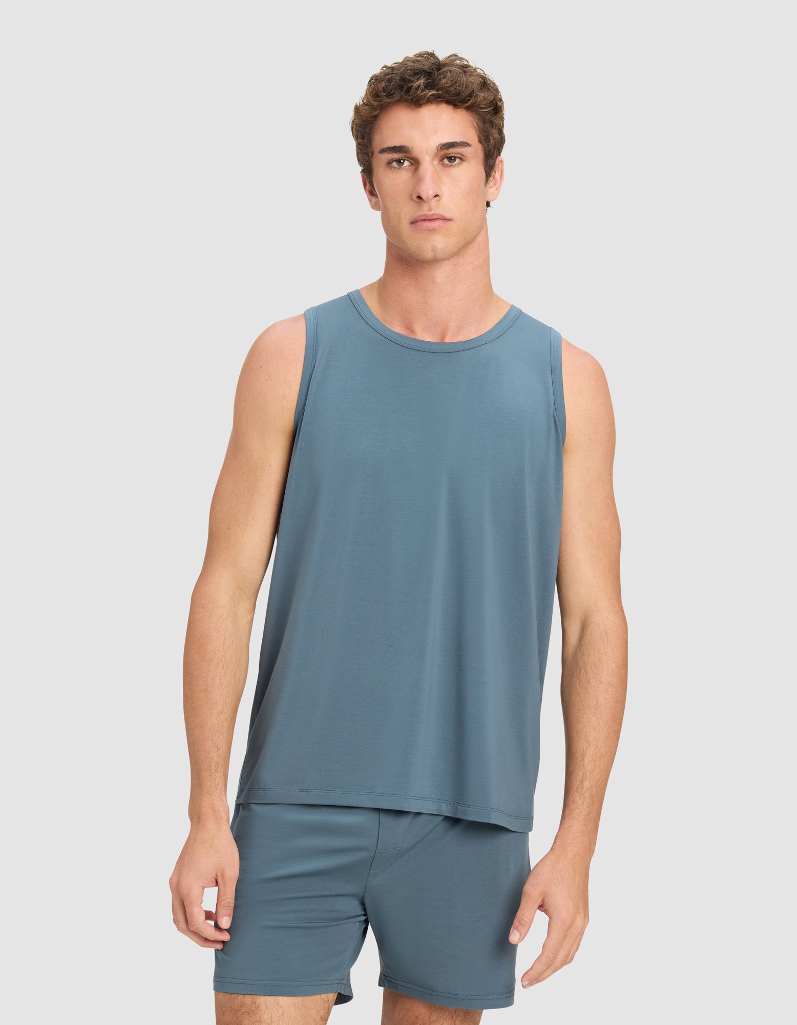 A young man with short, curly brown hair wears the Cozy Earth Men's First Layer Pajama Tank in blue, paired with matching shorts, standing against a plain light gray background and looking directly at the camera.