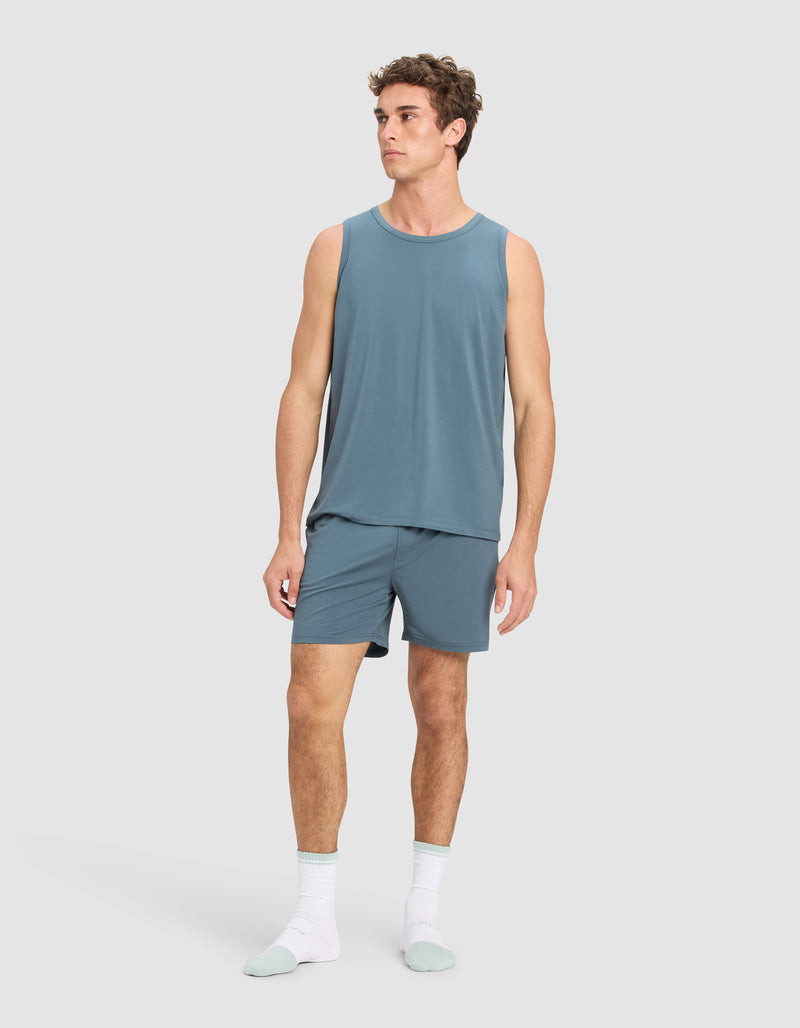A man stands against a plain background wearing the Cozy Earth Men's First Layer Pajama Tank and matching shorts, paired with white socks and light blue slippers. He looks slightly to his left with a neutral expression.