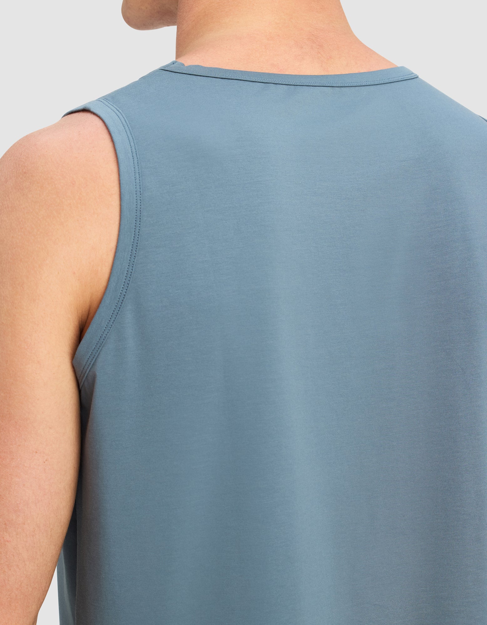 A close-up of a person wearing Cozy Earth's Men's First Layer Pajama Tank, sleeveless and blue, highlighting the upper back and shoulder against a plain light background.
