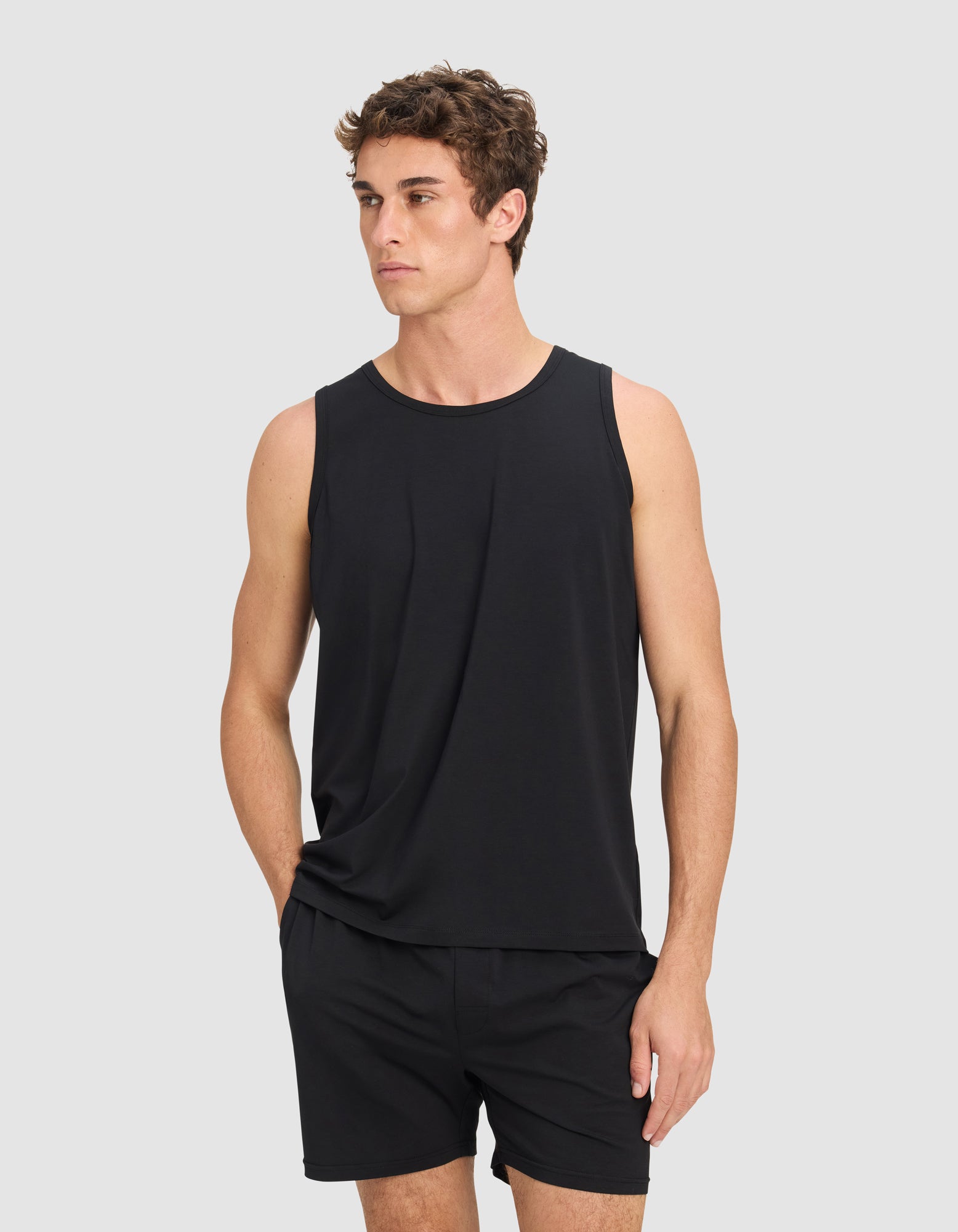A man wears the Men's First Layer Pajama Tank by Cozy Earth and black shorts, standing with one hand in his pocket against a plain light gray background.