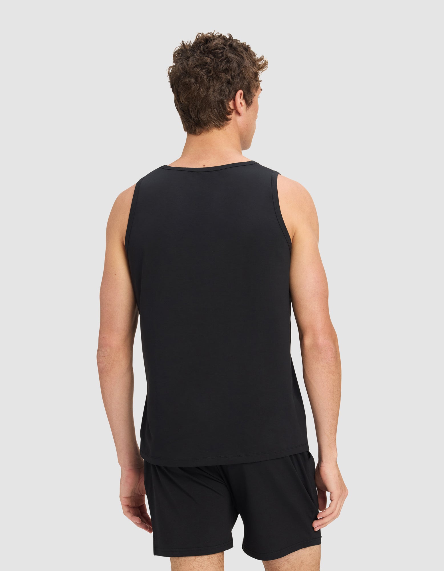 A man with short curly hair is seen from the back wearing Cozy Earth's Men's First Layer Pajama Tank in black, paired with black shorts, standing against a plain light gray background.|Color:Jet Black