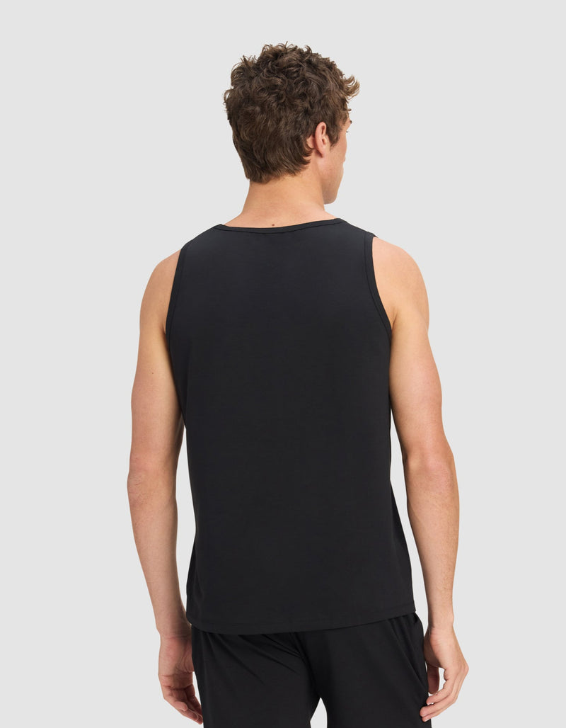 A man with short curly hair is seen from the back wearing Cozy Earth's Men's First Layer Pajama Tank in black, paired with black shorts, standing against a plain light gray background.