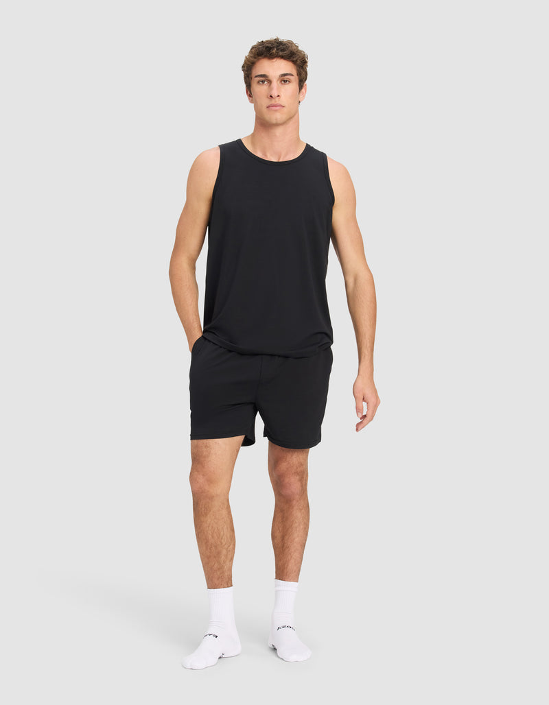 A man stands against a plain light gray background, wearing the Cozy Earth Men's First Layer Pajama Tank in black with black shorts, white socks, and white sneakers. His left hand is in his short pocket as he looks straight ahead.