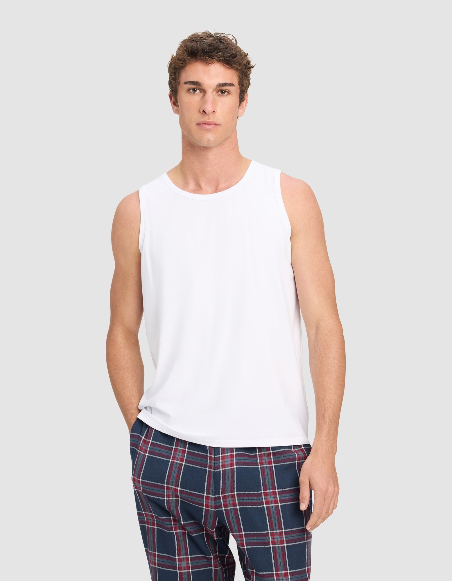 A man wears the Cozy Earth Men's First Layer Pajama Tank in white, paired with blue plaid pants, standing against a light gray background and looking directly at the camera with a neutral expression.|Color:White