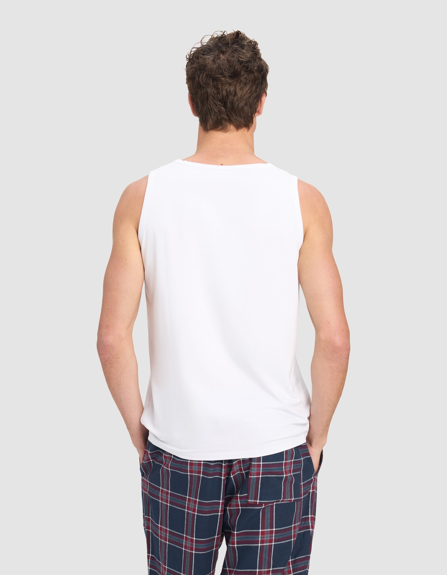 A man models the Cozy Earth Men's First Layer Pajama Tank in white with blue plaid pajama pants featuring red and white stripes, standing against a plain light gray background.