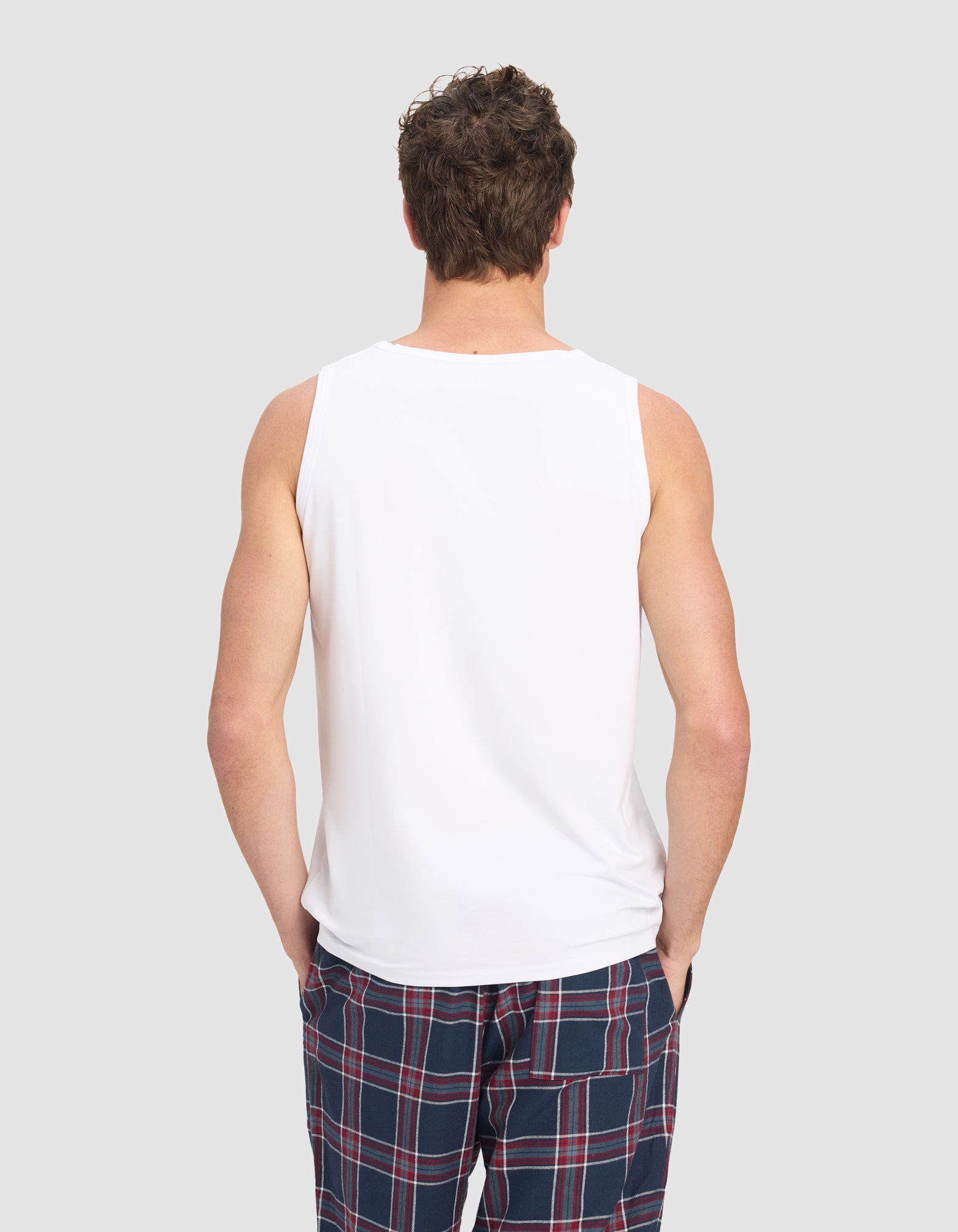 A man models the Cozy Earth Men's First Layer Pajama Tank in white with blue plaid pajama pants featuring red and white stripes, standing against a plain light gray background.