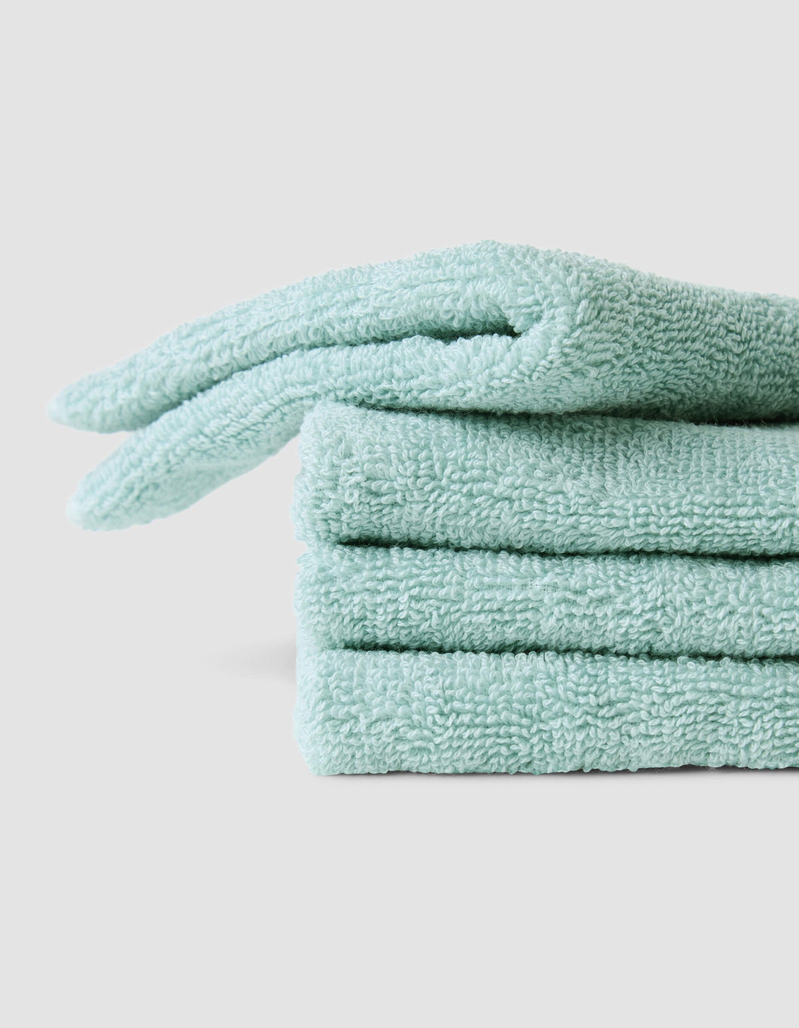 Three neatly folded Complete Cleansing Cloths by Cozy Earth, in light teal, are stacked on a plain light gray surface. The cloths have a soft, fluffy texture and appear clean and new.