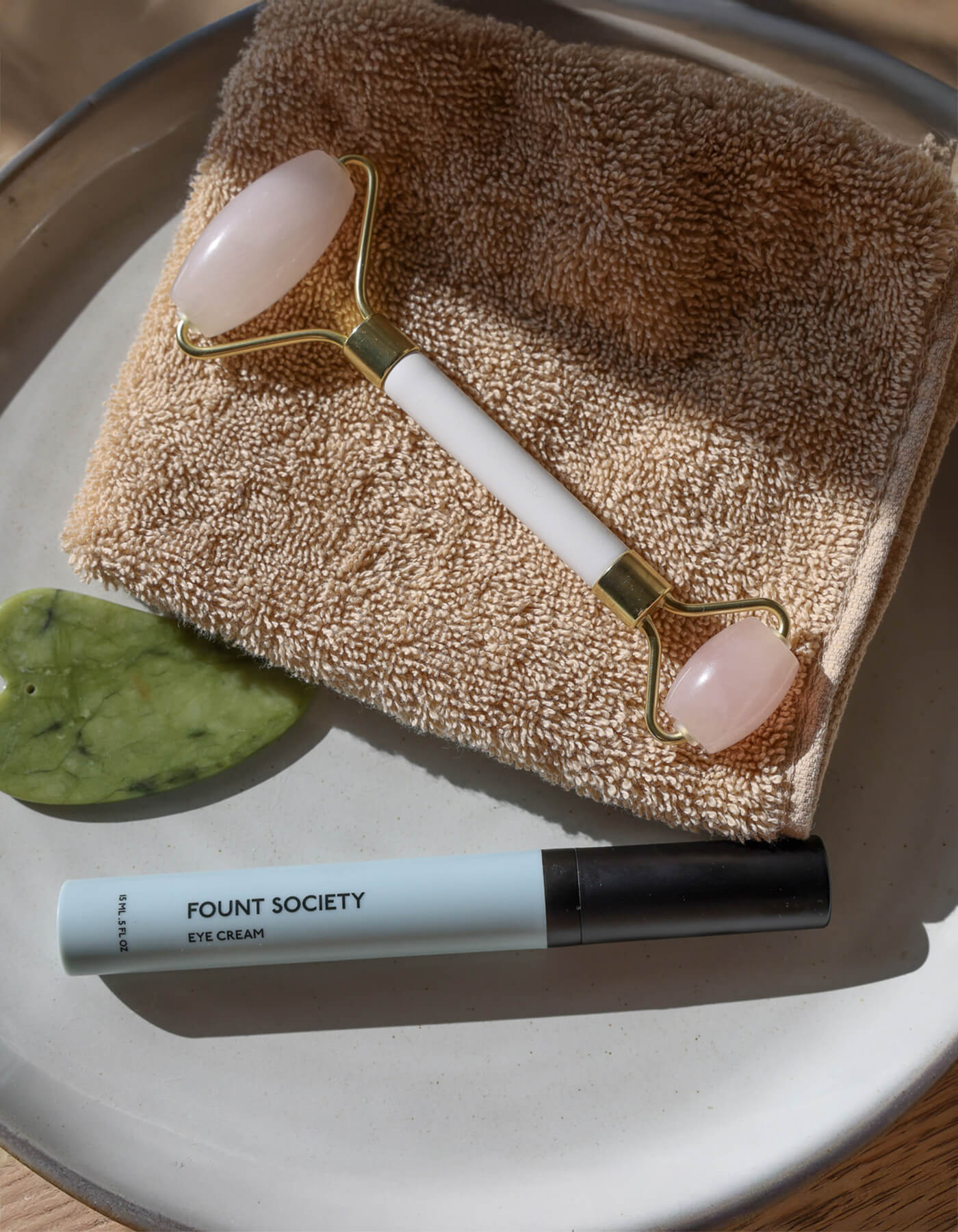 A jade roller, a gua sha stone, and a tube of Cozy Earth Eye Cream sit on a folded beige towel on a round plate, all illuminated by natural sunlight.