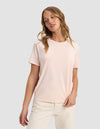 A woman with wavy blonde hair wears the Cozy Earth Women's Go-To Tee in Blossom and off-white pants, standing against a plain light gray background with a relaxed expression, looking slightly to the side.|Color:Blossom