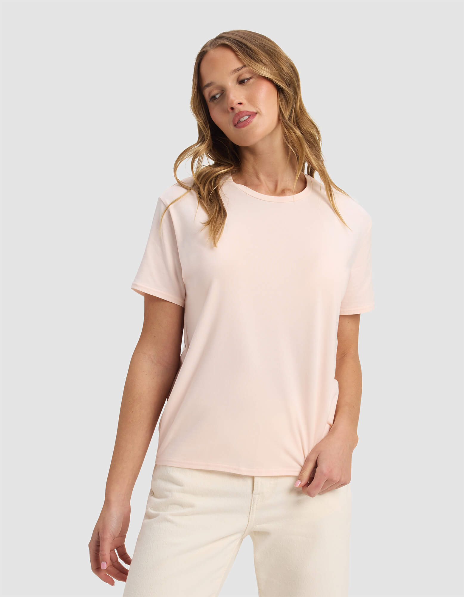 A woman with wavy blonde hair wears the Cozy Earth Women's Go-To Tee in Blossom and off-white pants, standing against a plain light gray background with a relaxed expression, looking slightly to the side.