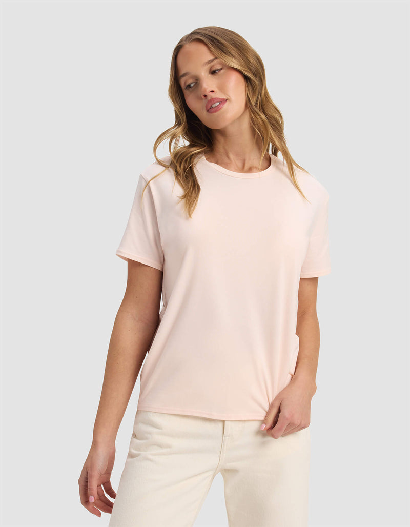 A woman with wavy blonde hair wears the Cozy Earth Women's Go-To Tee in Blossom and off-white pants, standing against a plain light gray background with a relaxed expression, looking slightly to the side.