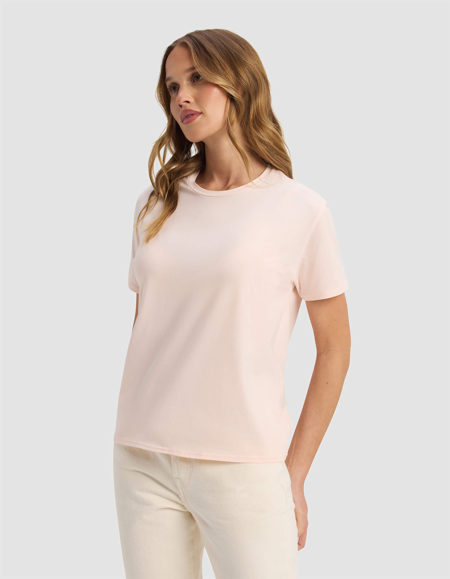 A woman with wavy light brown hair wears the Cozy Earth Women's Go-To Tee in pale pink and off-white pants, standing against a plain light gray background and gazing to the side with a relaxed expression.