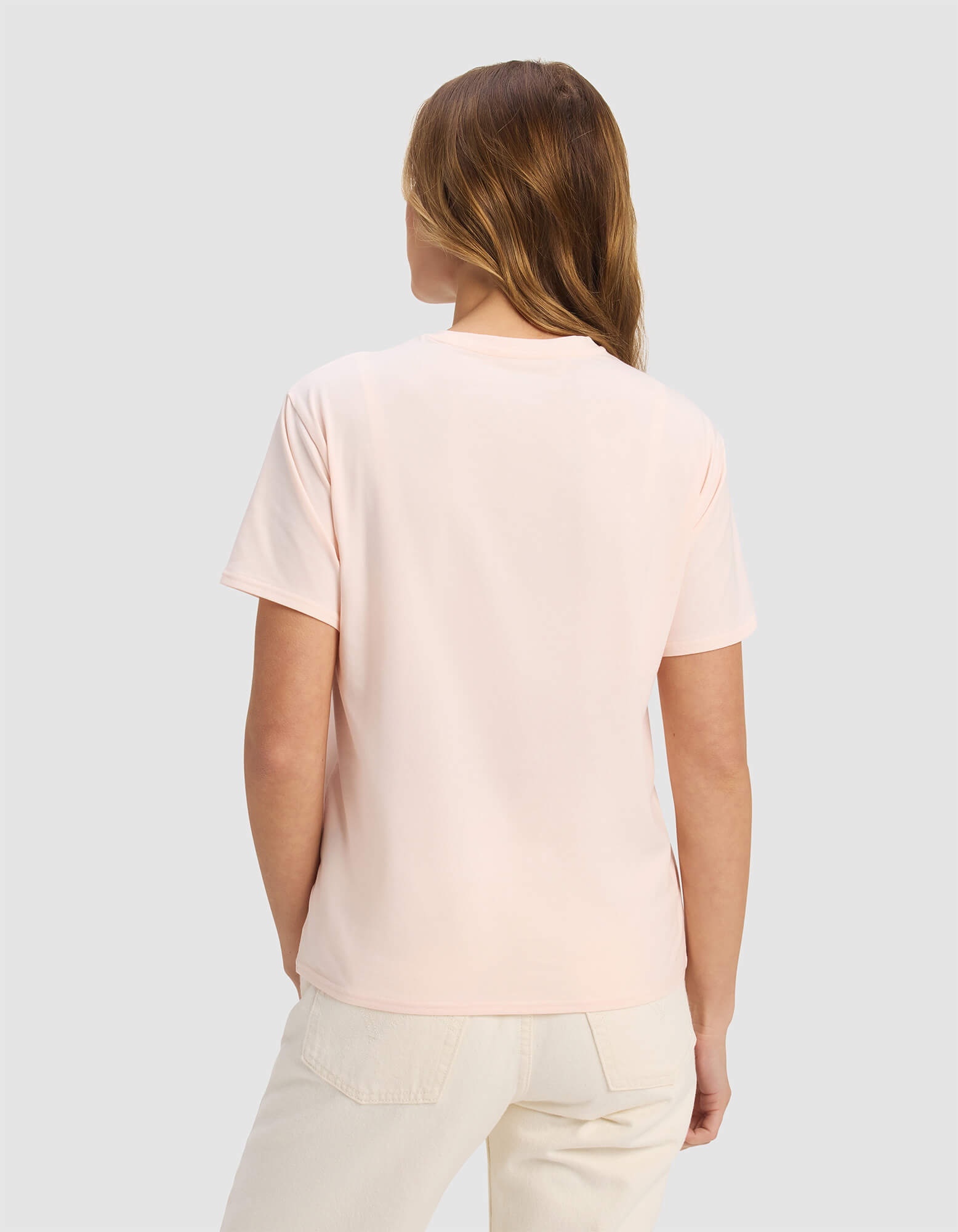 A woman with light brown hair stands with her back to the camera, wearing a Cozy Earth Women's Go-To Tee in pale pink and light-colored pants against a plain light gray background.|Color:Blossom